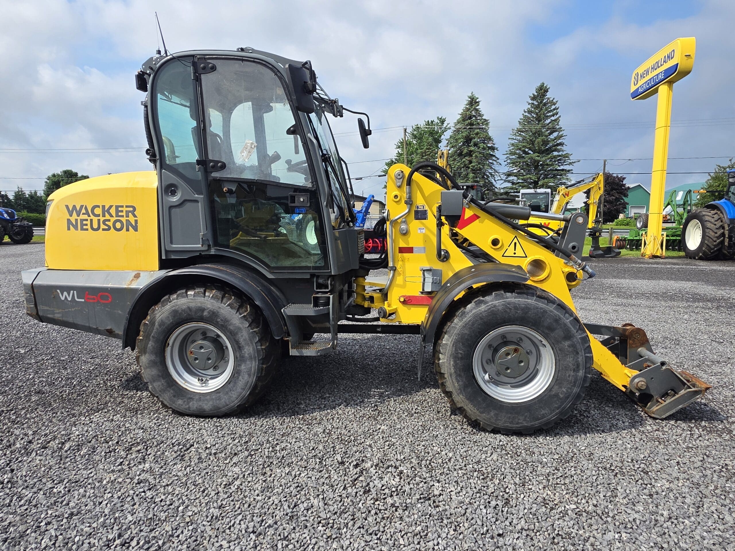 Une chargeuse compacte sur pneus Wacker Neuson WL50 jaune et noire est garée sur du gravier près d'un concessionnaire, avec des arbres et des tracteurs visibles à l'arrière-plan.