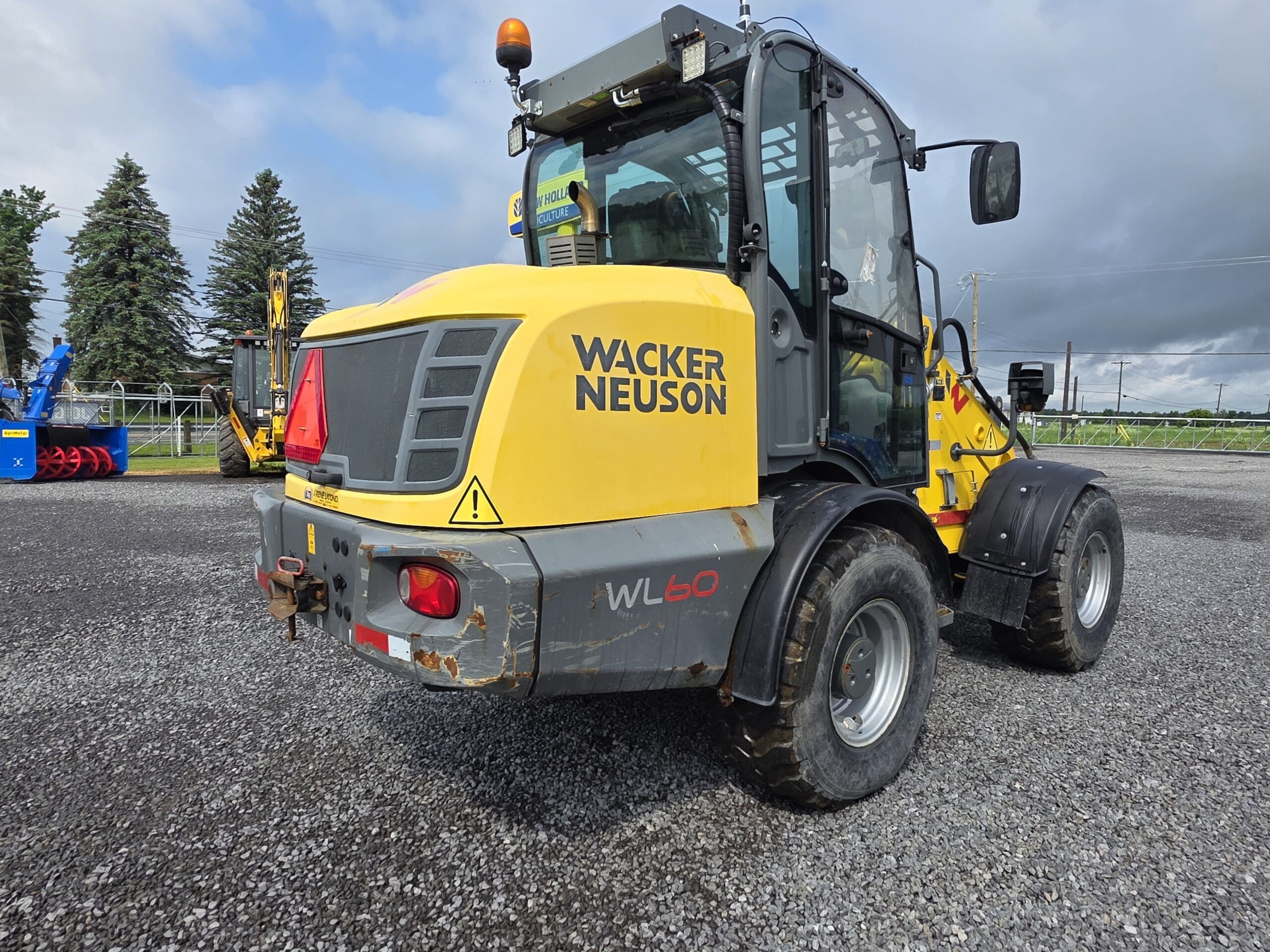 Une chargeuse sur pneus Wacker Neuson WL60 jaune est garée sur une surface de gravier en plein air, avec des arbres et d'autres machines visibles en arrière-plan sous un ciel nuageux.