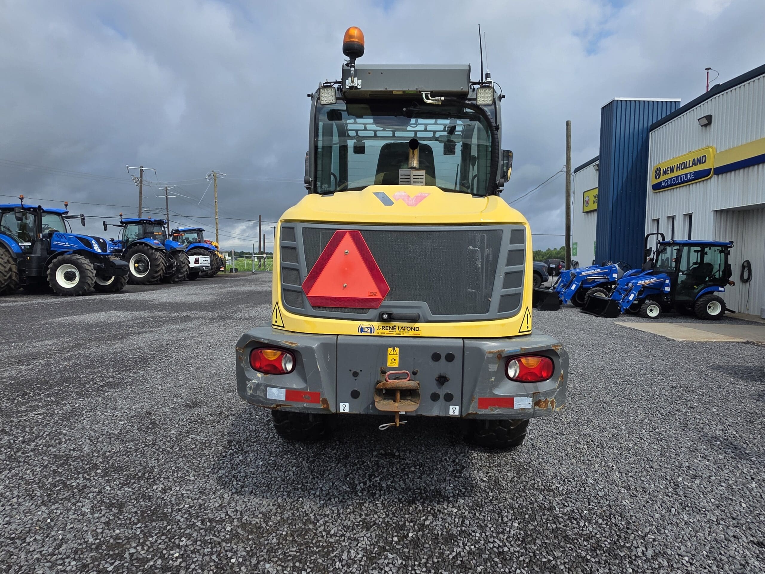 Vue arrière d'un véhicule de chantier jaune avec un panneau triangulaire rouge, garé sur un terrain en gravier près d'un bâtiment et de plusieurs tracteurs bleus sous un ciel nuageux.