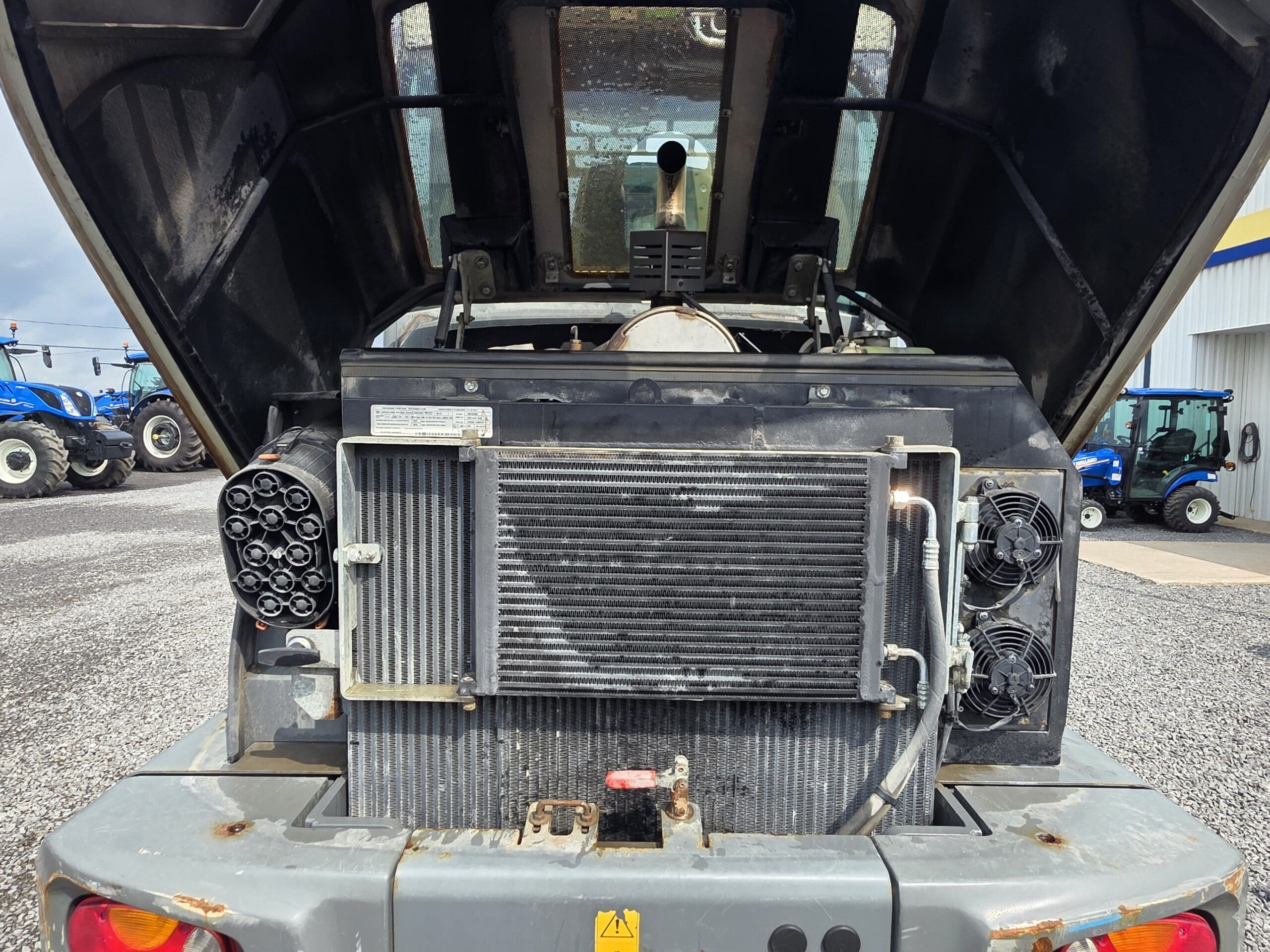 Vue du compartiment moteur d'un équipement lourd avec le capot ouvert, montrant le radiateur, les ventilateurs de refroidissement, les tuyaux et les composants du moteur. D'autres machines agricoles sont visibles à l'arrière-plan, à l'extérieur, sur un terrain en gravier.