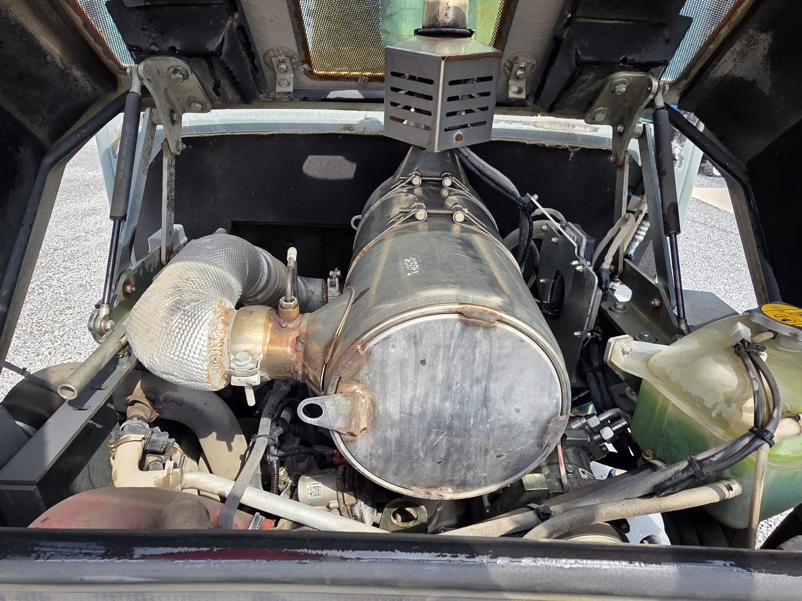 Vue rapprochée du compartiment moteur d'un véhicule avec divers composants en métal et en caoutchouc, y compris une pièce cylindrique avec des boulons, des tuyaux isolés et un réservoir de liquide de refroidissement translucide sur la droite.