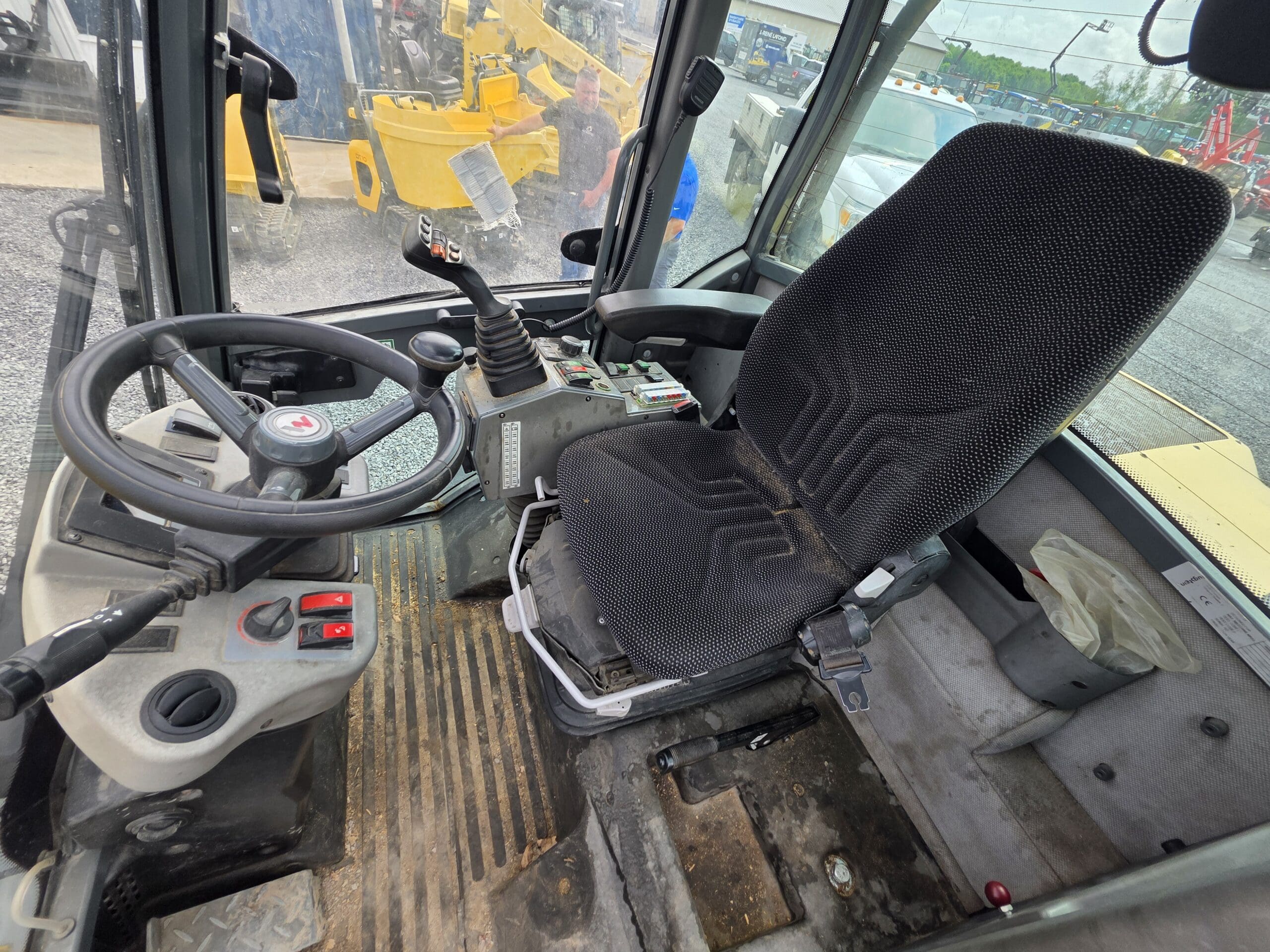 L'image montre l'intérieur de la cabine d'un véhicule de construction, avec un siège de conducteur noir, un volant, des leviers de commande, des pédales et un sol sale. Différents interrupteurs et leviers de commande sont situés autour du siège.