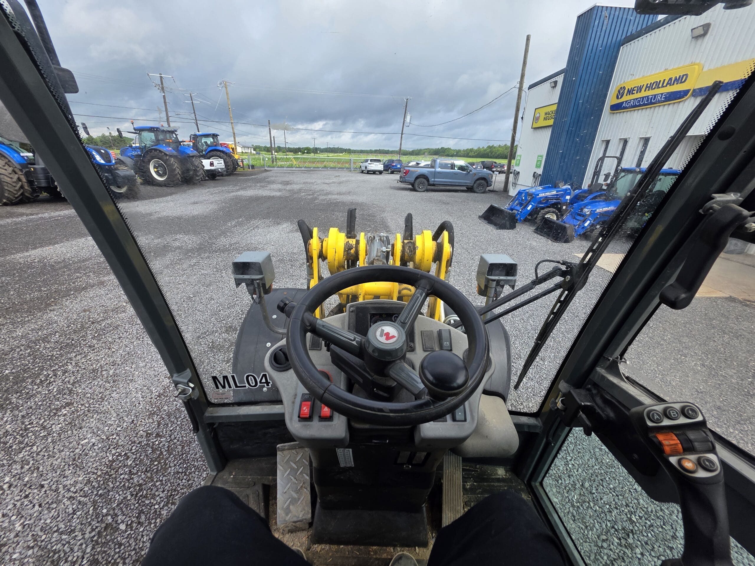 Vue de l'intérieur d'une cabine de chargeur montrant le volant, les commandes et les bras jaunes du chargeur. À l'extérieur, des tracteurs, des voitures et un bâtiment avec des panneaux New Holland sur un terrain en gravier sous un ciel nuageux.