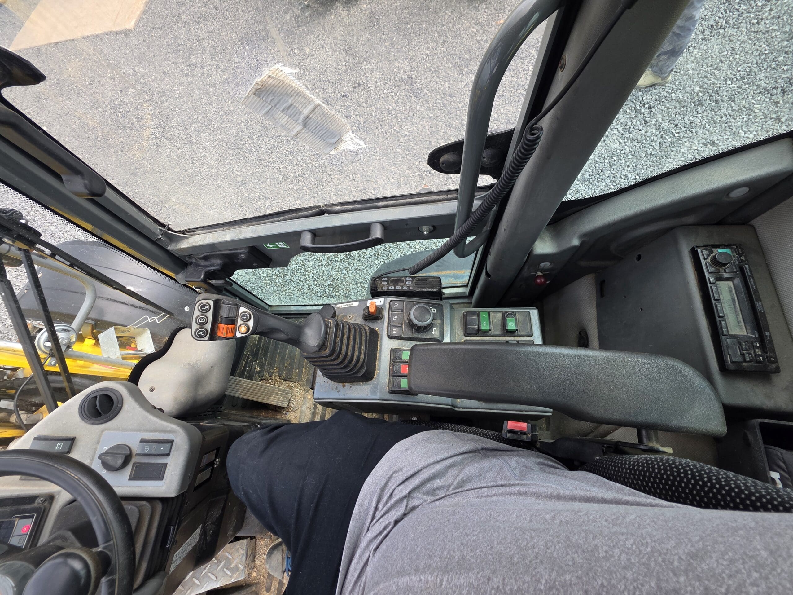 Une personne est assise dans la cabine d'un engin de chantier, près du panneau de commande comportant des boutons, des leviers et un joystick, avec une vue sur le sol et le gravier à l'extérieur par la fenêtre.