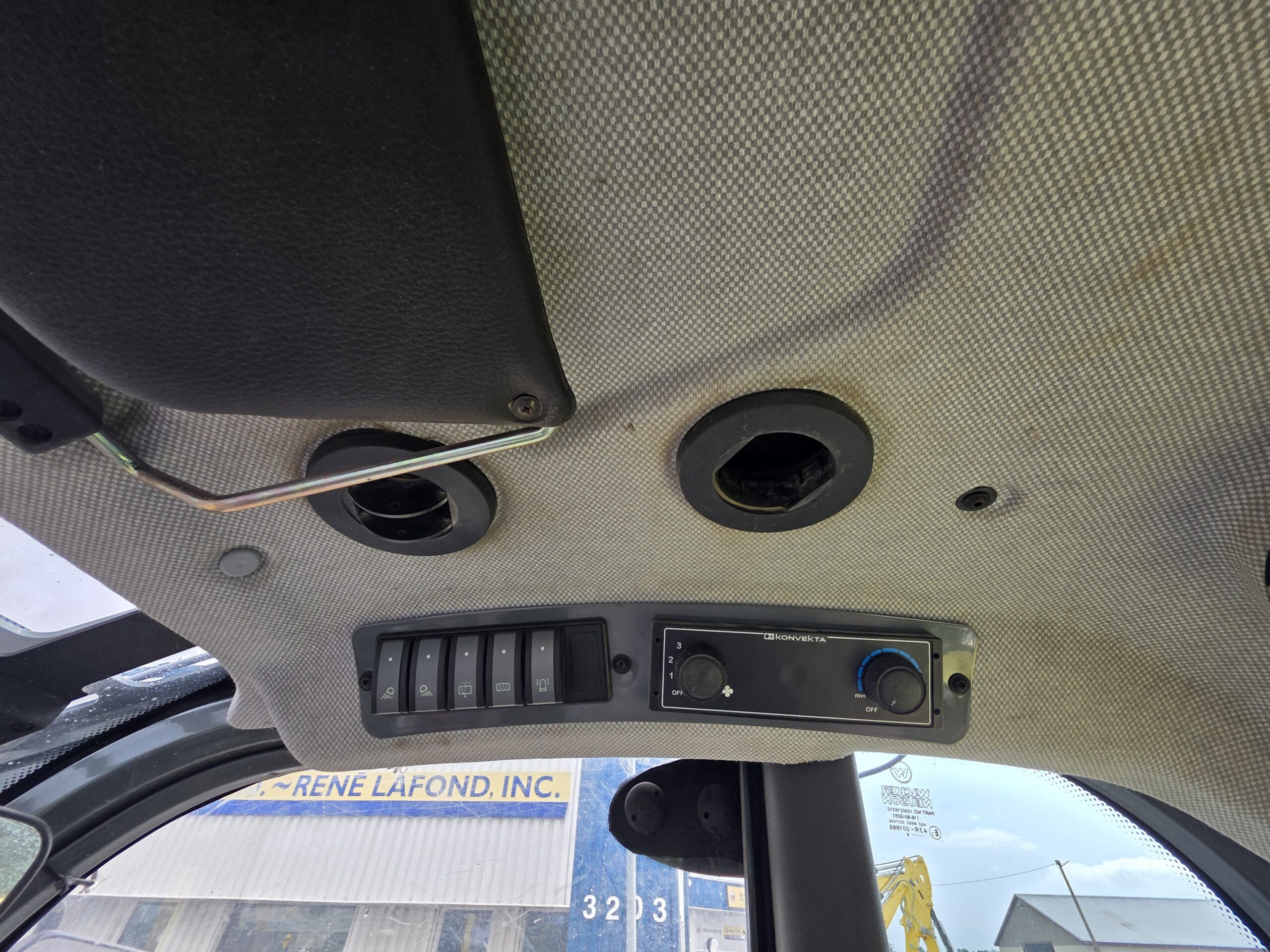 Vue intérieure du panneau de plafond d'un véhicule avec des bouches d'aération rondes, une rangée d'interrupteurs à bascule et un module de commande. Une partie d'une fenêtre et un panneau de signalisation extérieur sont visibles à travers le pare-brise.