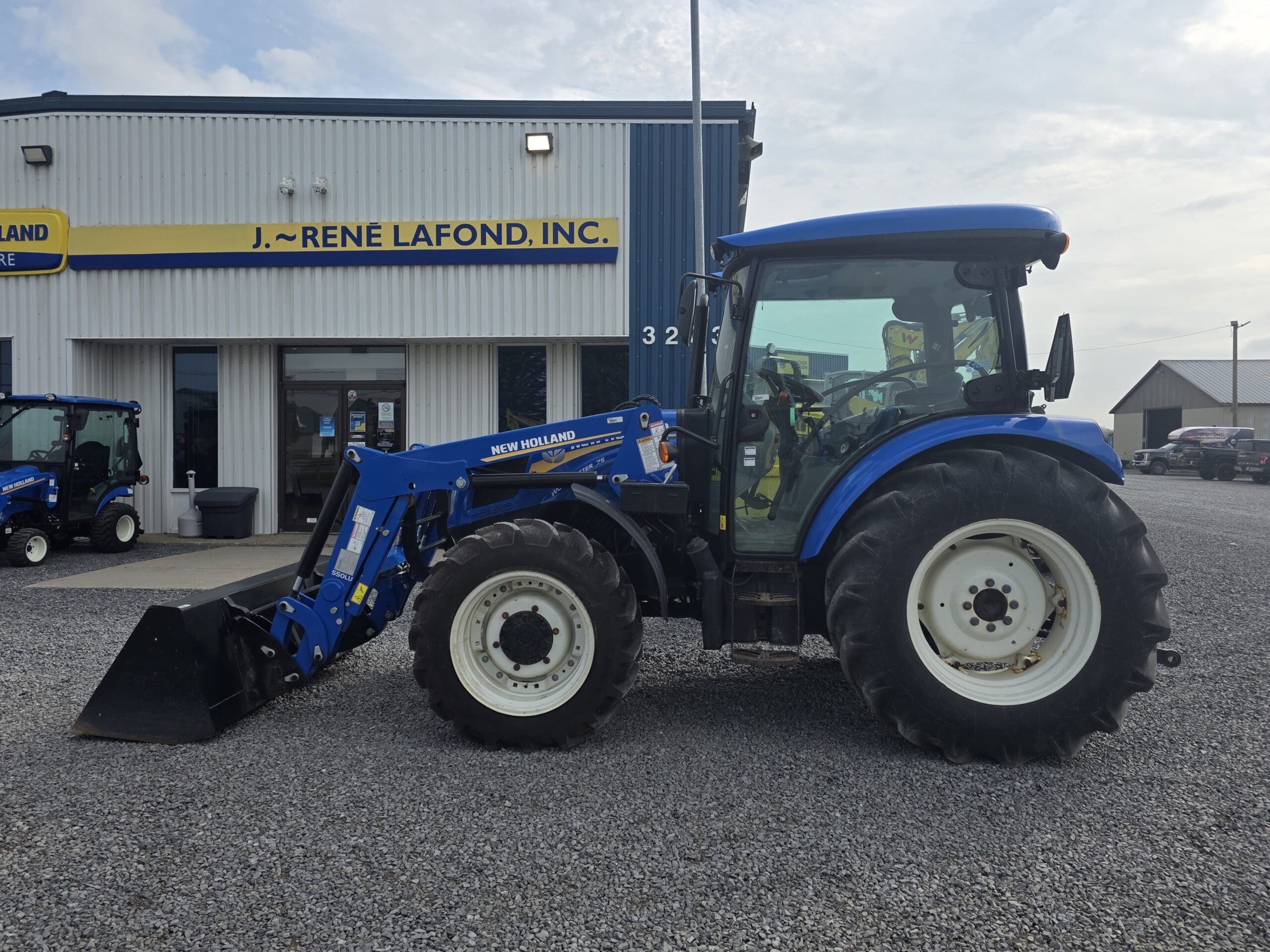 Par une journée nuageuse, un tracteur New Holland bleu équipé d'un chargeur frontal est garé sur du gravier devant un bâtiment portant l'inscription