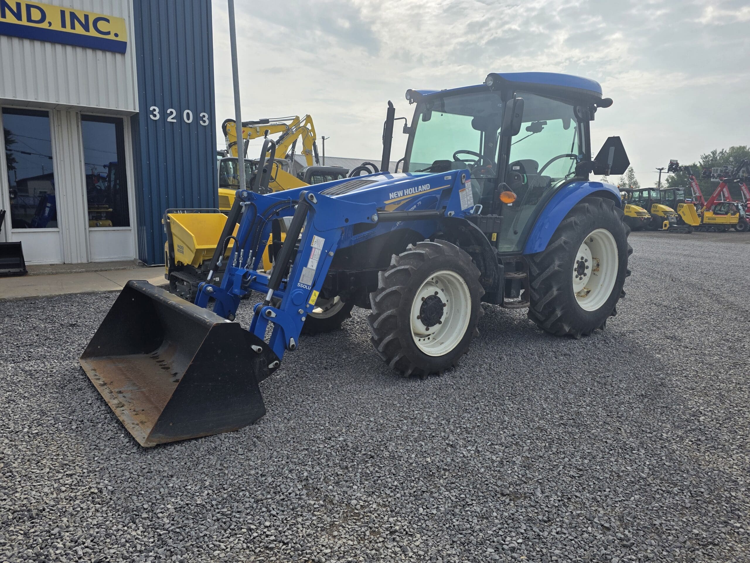 Un tracteur New Holland bleu équipé d'un chargeur frontal est garé sur du gravier à l'extérieur d'un bâtiment bleu et blanc portant le numéro 3203. D'autres machines jaunes sont visibles à l'arrière-plan.
