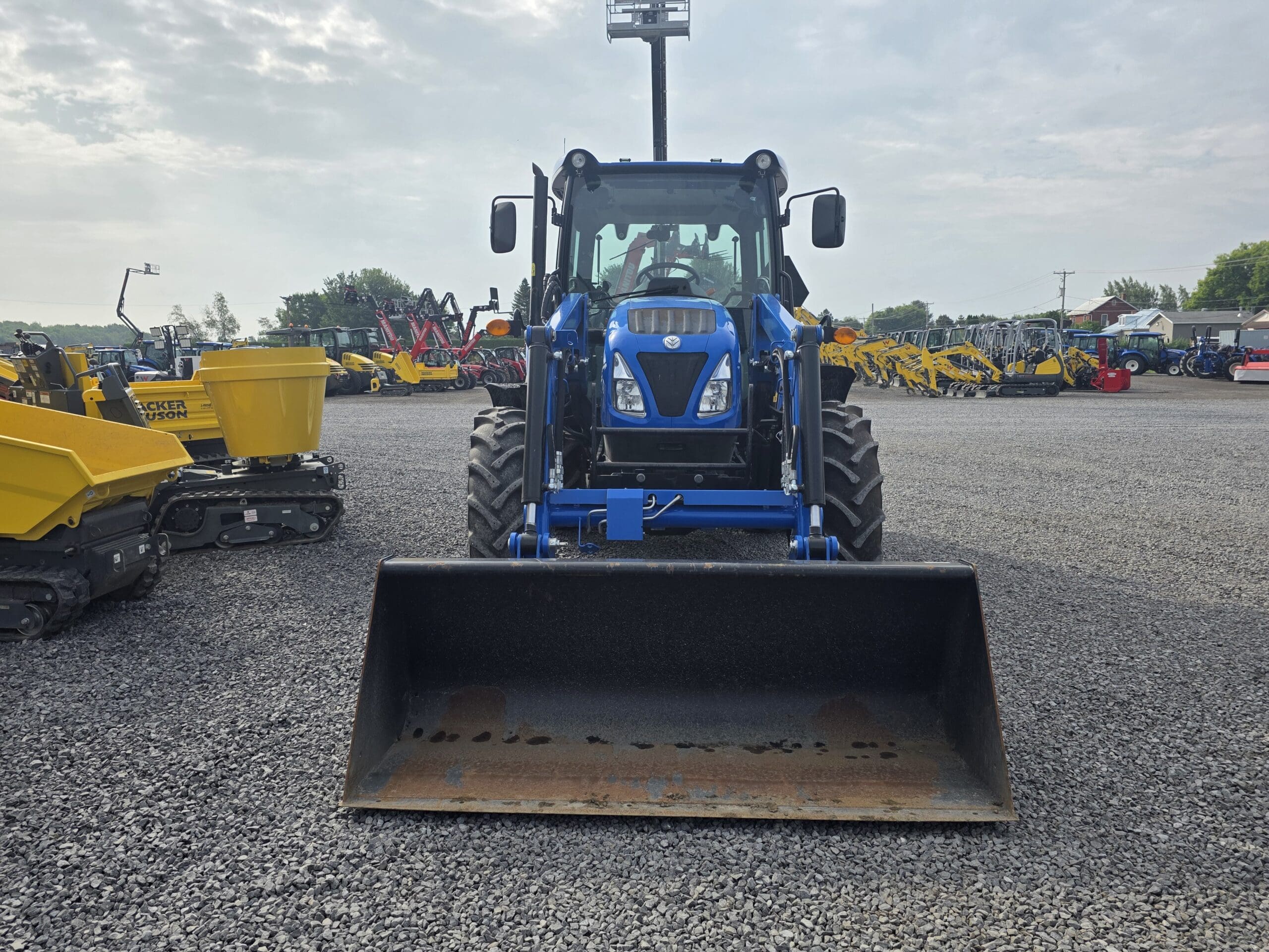 Un tracteur bleu à chargeur frontal est garé sur un terrain en gravier avec d'autres véhicules de construction et agricoles en arrière-plan sous un ciel partiellement nuageux.