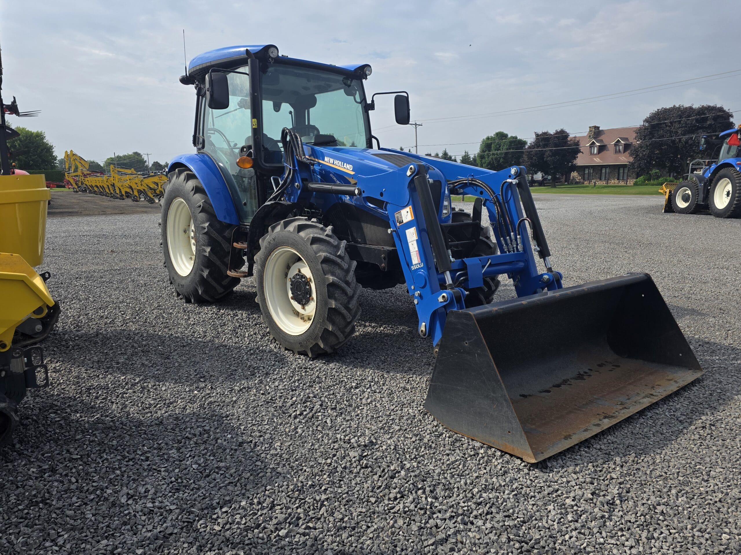 Un tracteur New Holland bleu équipé d'un chargeur frontal est garé sur un terrain en gravier sous un ciel partiellement nuageux. D'autres équipements et bâtiments agricoles sont visibles à l'arrière-plan.