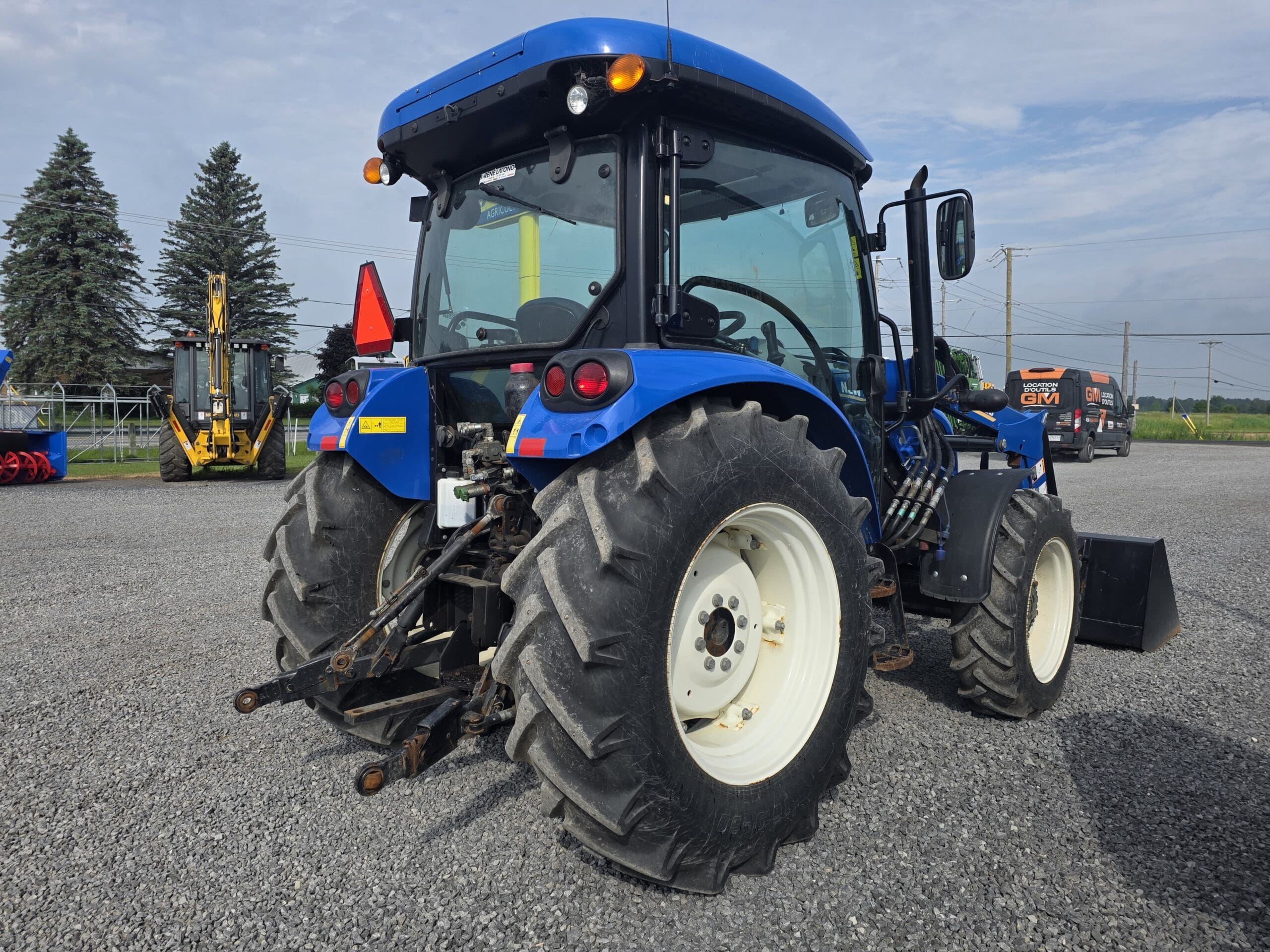 Un tracteur bleu avec de gros pneus arrière garé sur un terrain en gravier, avec du matériel agricole et des champs verts visibles en arrière-plan sous un ciel partiellement nuageux.