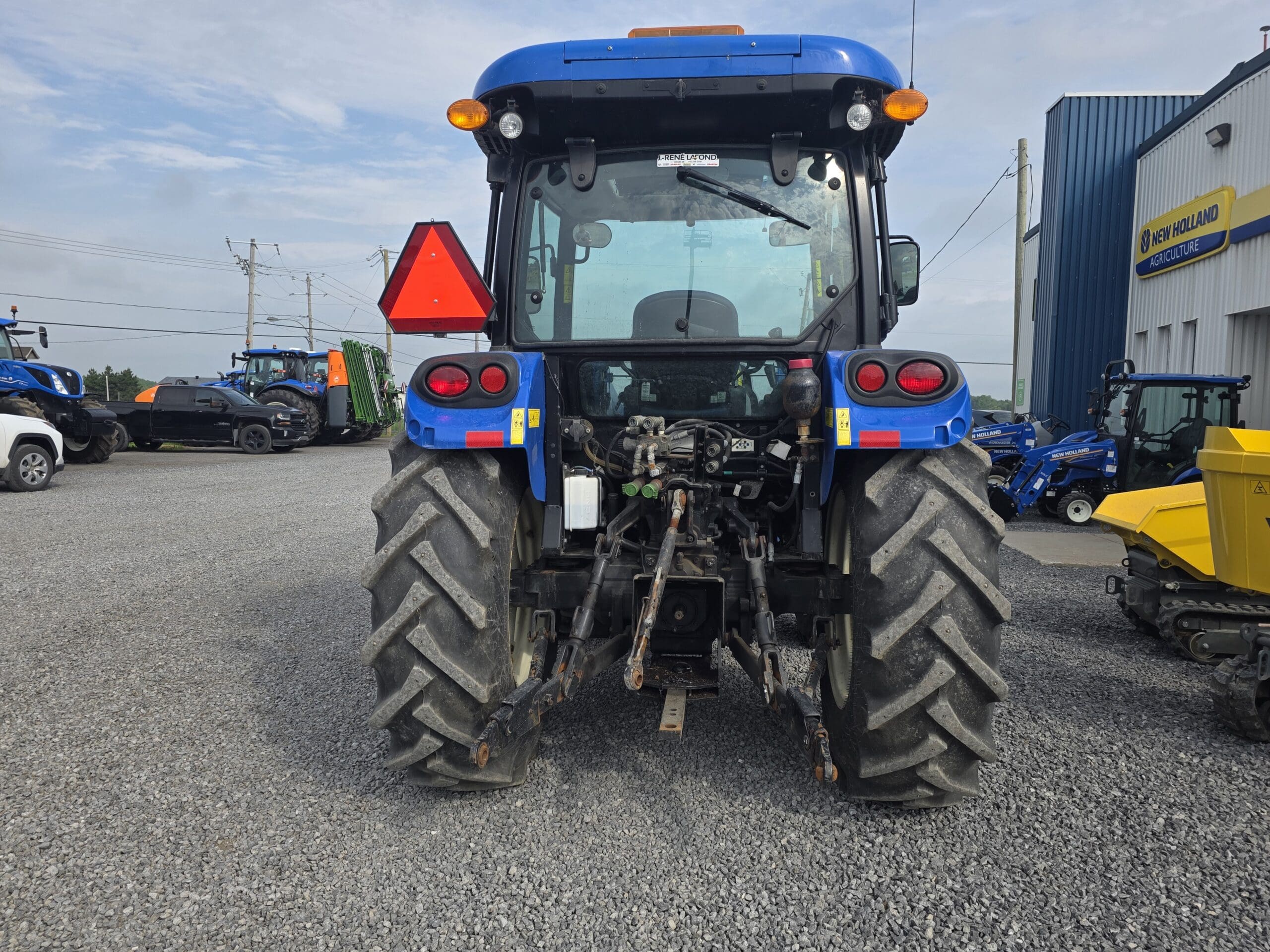 Un tracteur bleu, vu de l'arrière, est garé sur un terrain en gravier, avec un triangle d'avertissement rouge attaché. D'autres équipements et véhicules sont visibles à l'arrière-plan, près d'un bâtiment bleu et jaune de la concession New Holland.