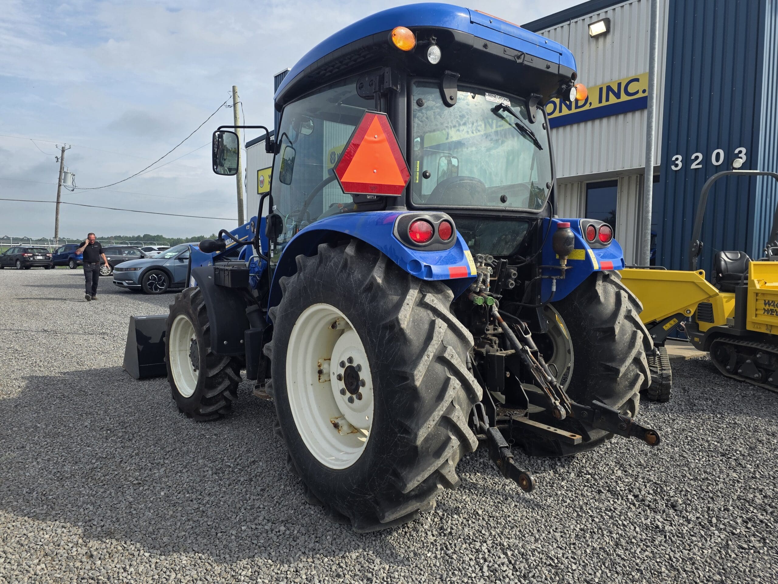 Un tracteur bleu avec de gros pneus arrière est garé sur du gravier à l'extérieur d'un bâtiment. Un panneau rouge et orange indiquant qu'il s'agit d'un véhicule lent est placé à l'arrière. D'autres véhicules et une personne marchant sont visibles à l'arrière-plan.