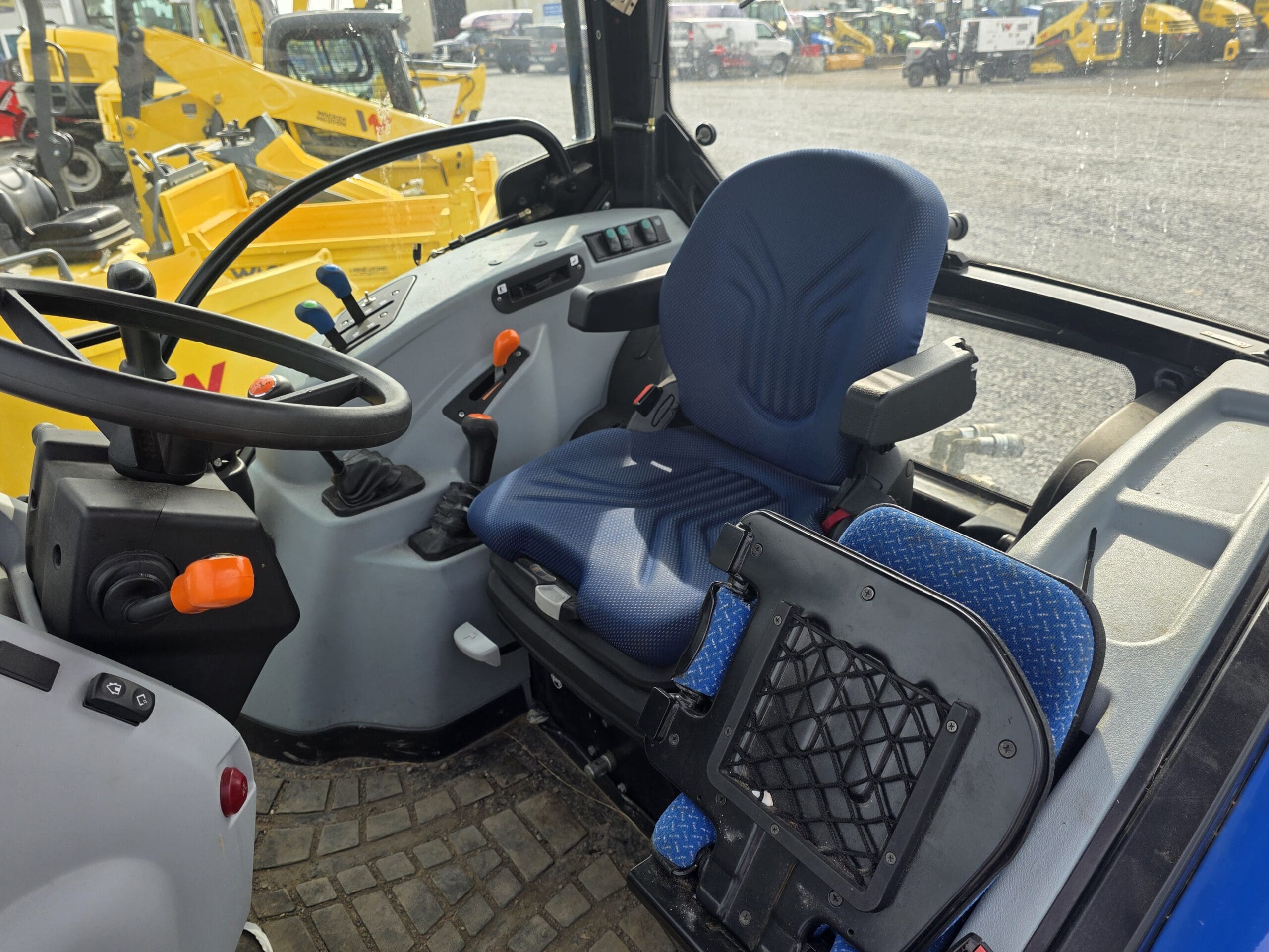 L'image montre le siège du conducteur et le panneau de commande à l'intérieur de la cabine d'un tracteur, avec un siège à coussin bleu, un volant, divers leviers et boutons. Des véhicules de construction jaunes sont visibles à travers les fenêtres à l'extérieur.