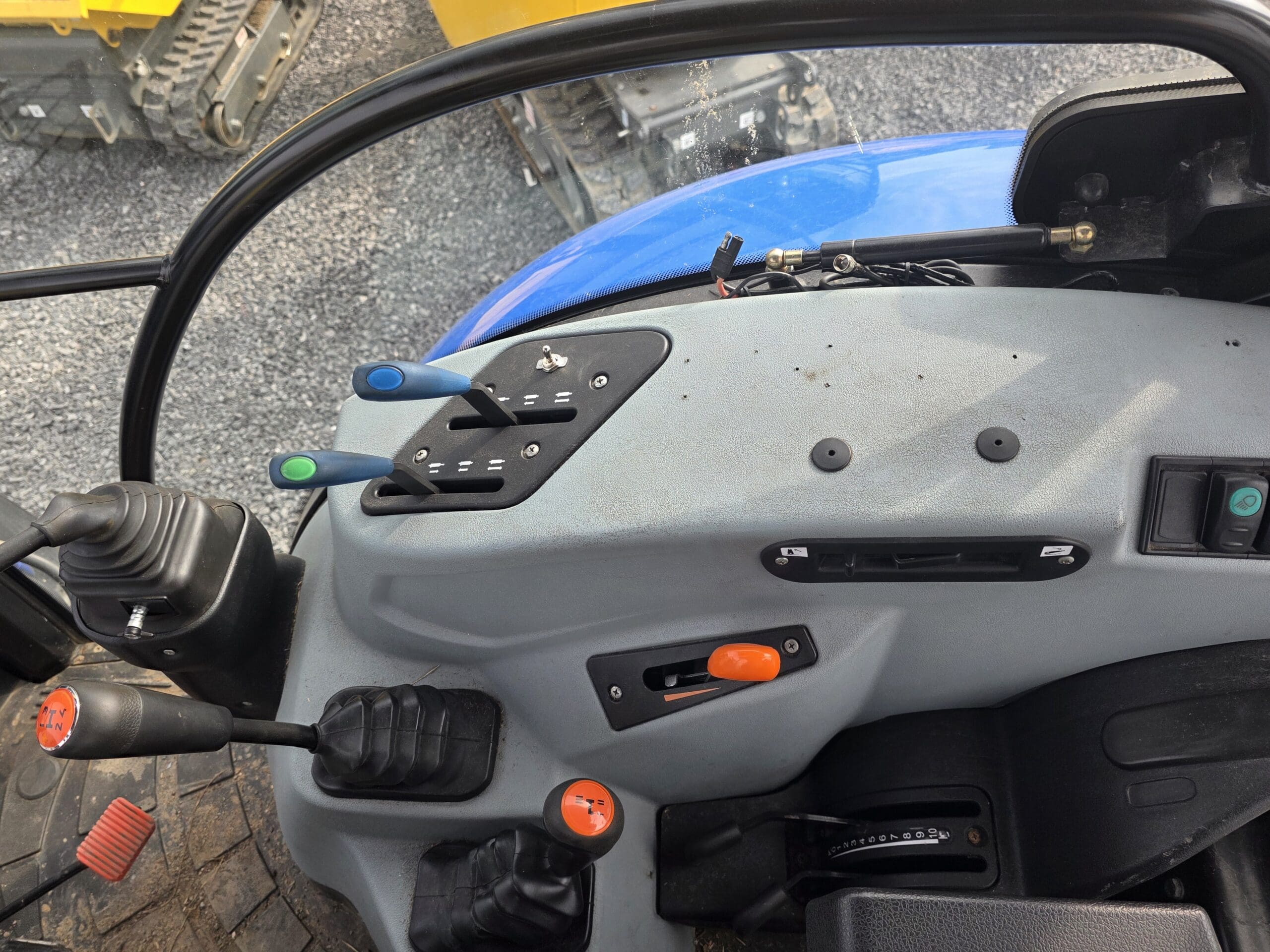 Vue rapprochée du panneau de commande à l'intérieur d'un tracteur, montrant des leviers, des interrupteurs, des pédales et des boutons sur un tableau de bord gris et noir. L'extérieur d'un engin de chantier est visible par la fenêtre.