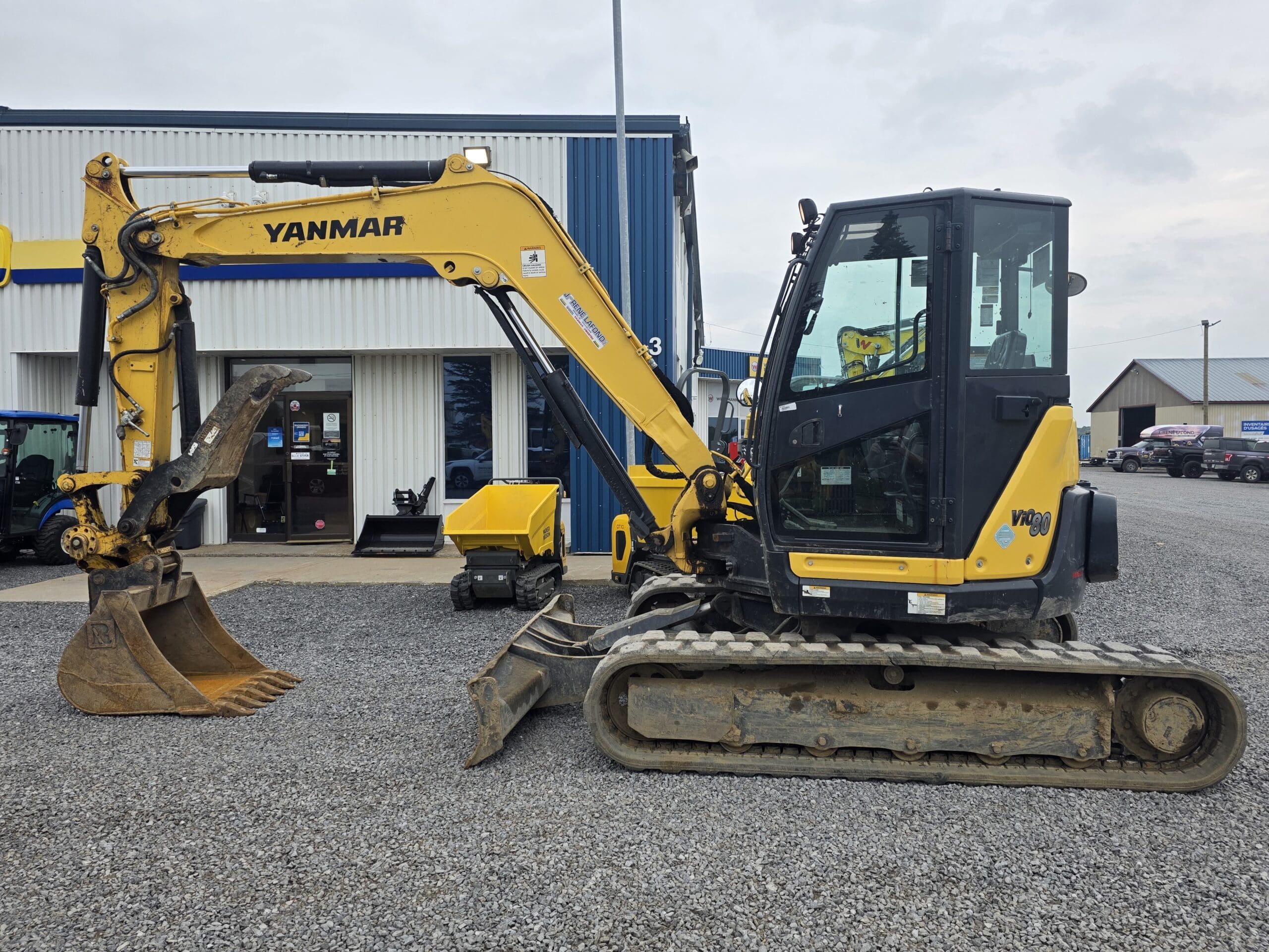 Pelle compacte Yanmar jaune garée sur du gravier à l'extérieur d'un bâtiment avec un revêtement bleu et blanc et une porte vitrée. La machine est équipée d'un godet d'excavation et est positionnée latéralement.