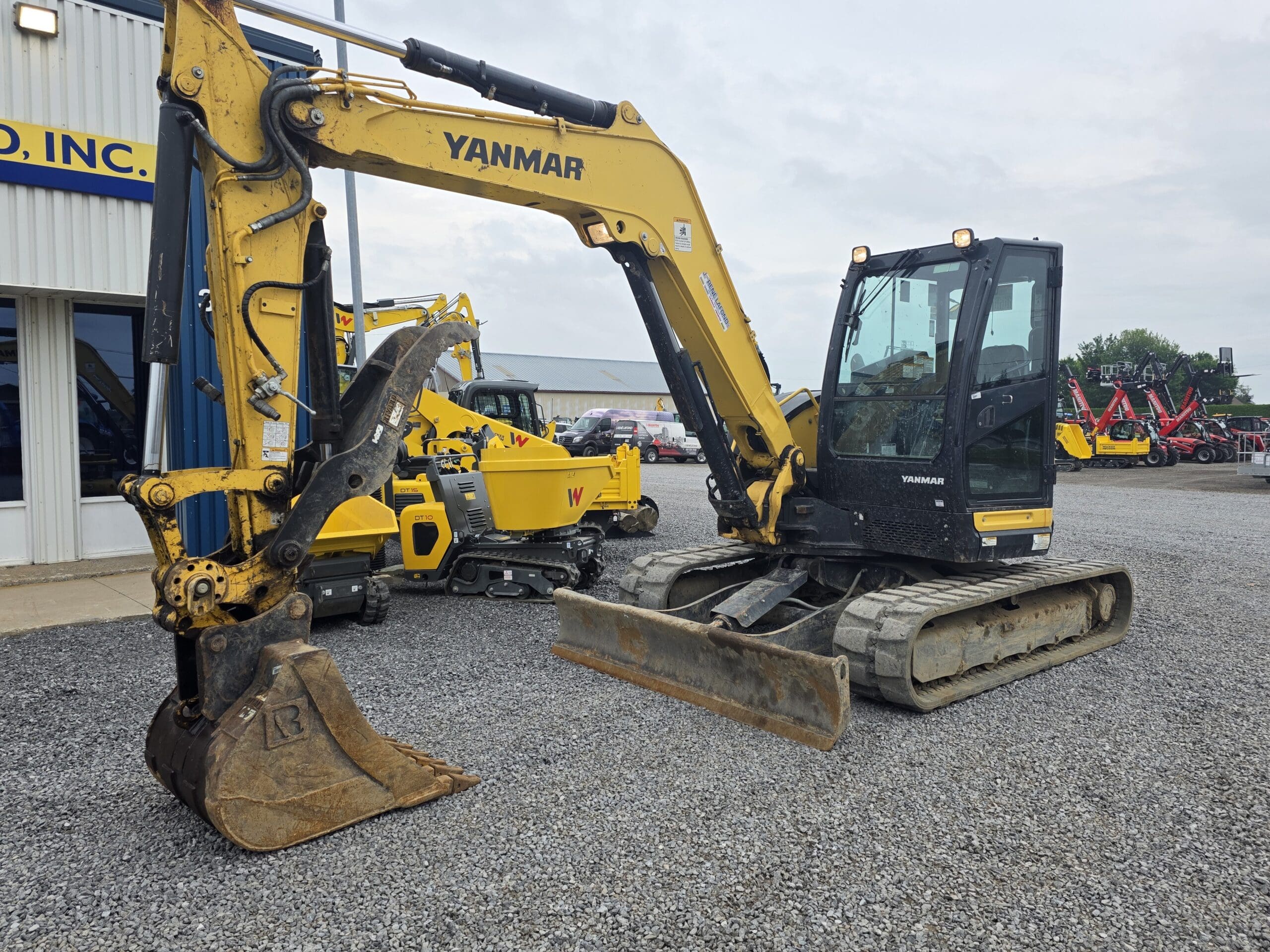 Une pelleteuse Yanmar jaune équipée d'un godet métallique est garée sur du gravier à l'extérieur d'un bâtiment industriel. D'autres engins de chantier sont visibles à l'arrière-plan.