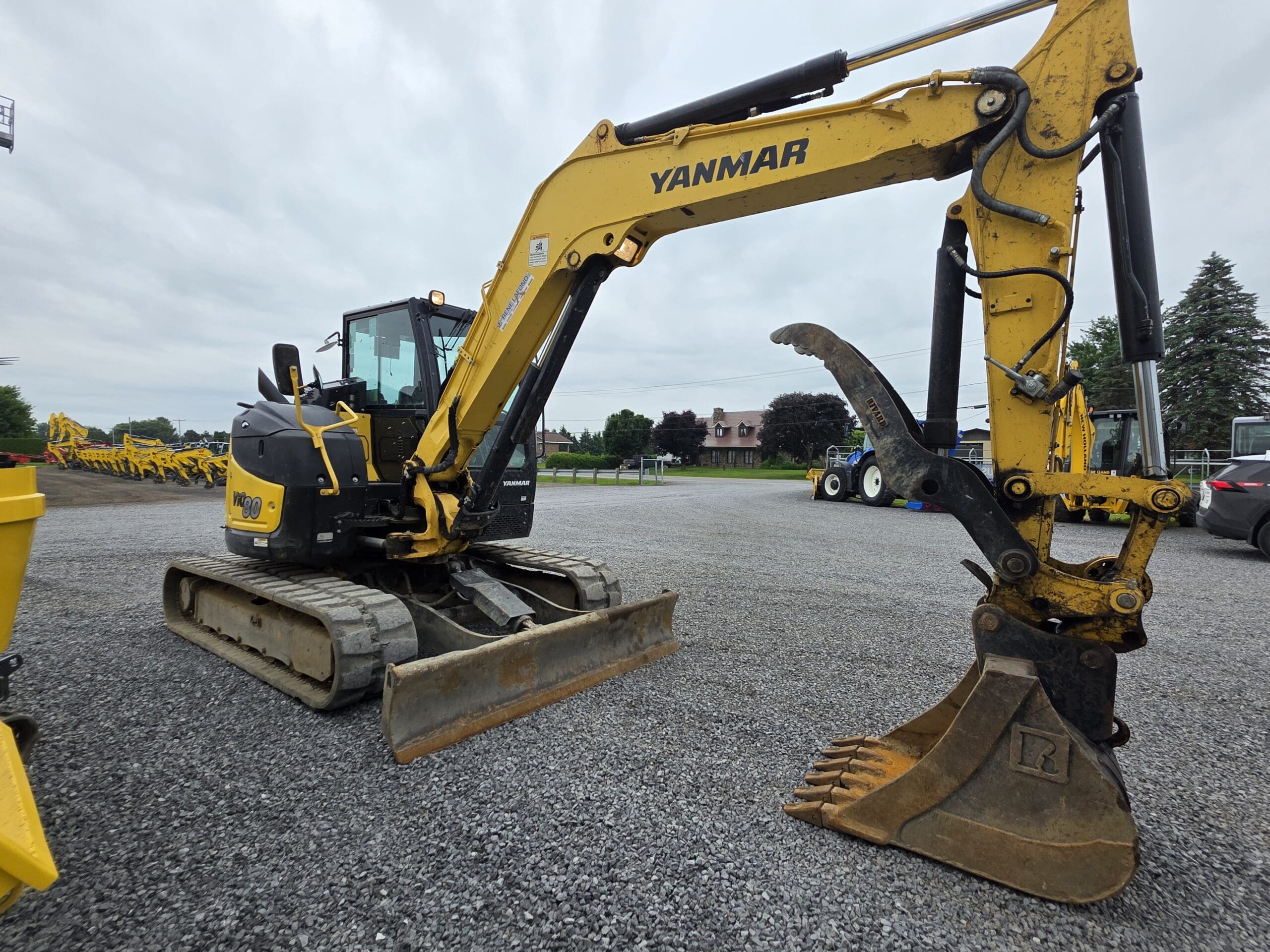 Une pelleteuse Yanmar jaune équipée d'une pince hydraulique est garée sur du gravier, avec plusieurs autres véhicules de construction en arrière-plan, sous un ciel nuageux.