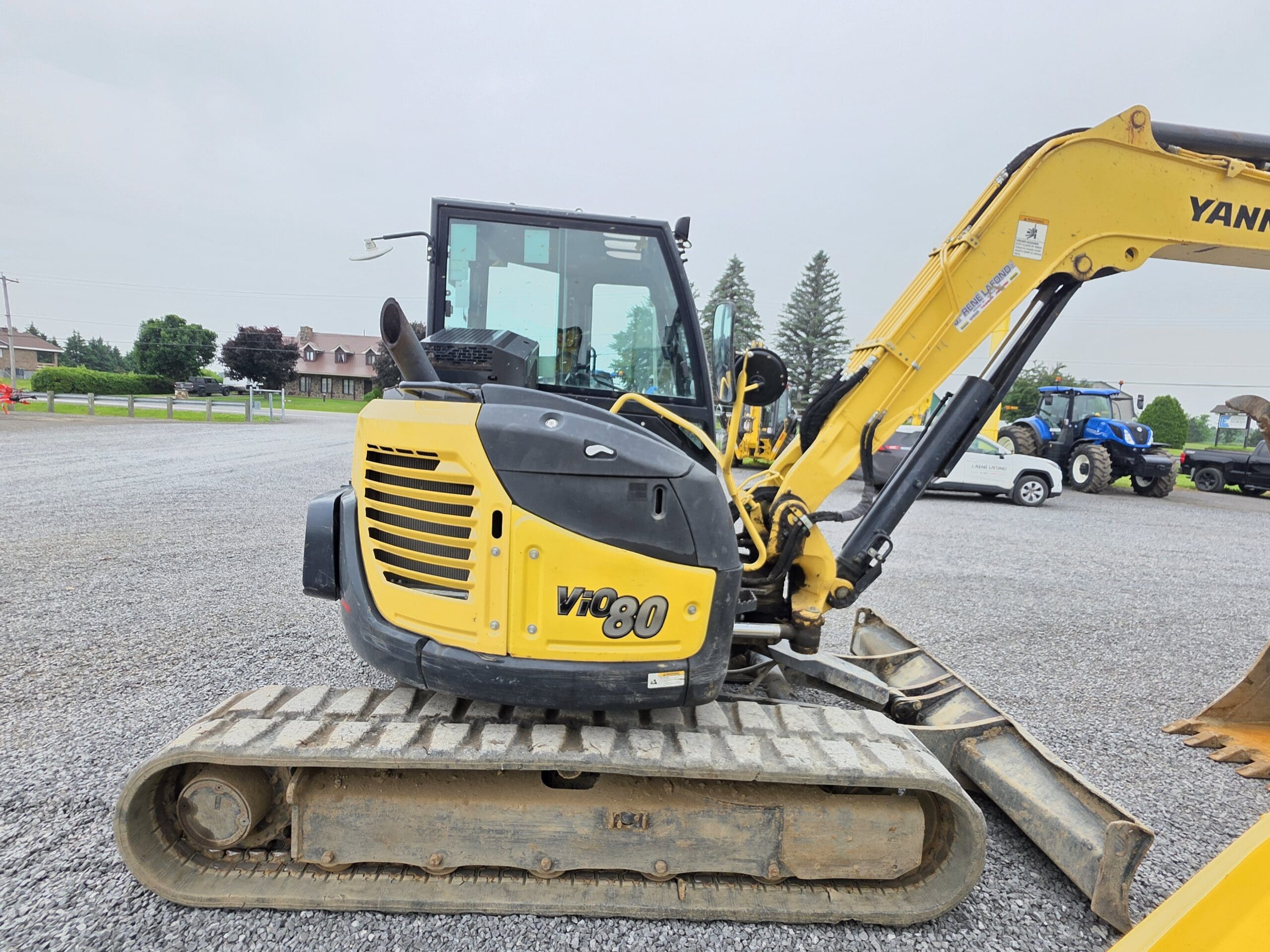 Une excavatrice compacte Yanmar VIO80 jaune et noire est posée sur un terrain en gravier, avec sa cabine et son bras visibles. Des arbres, des bâtiments et d'autres véhicules se trouvent à l'arrière-plan, sous un ciel nuageux.