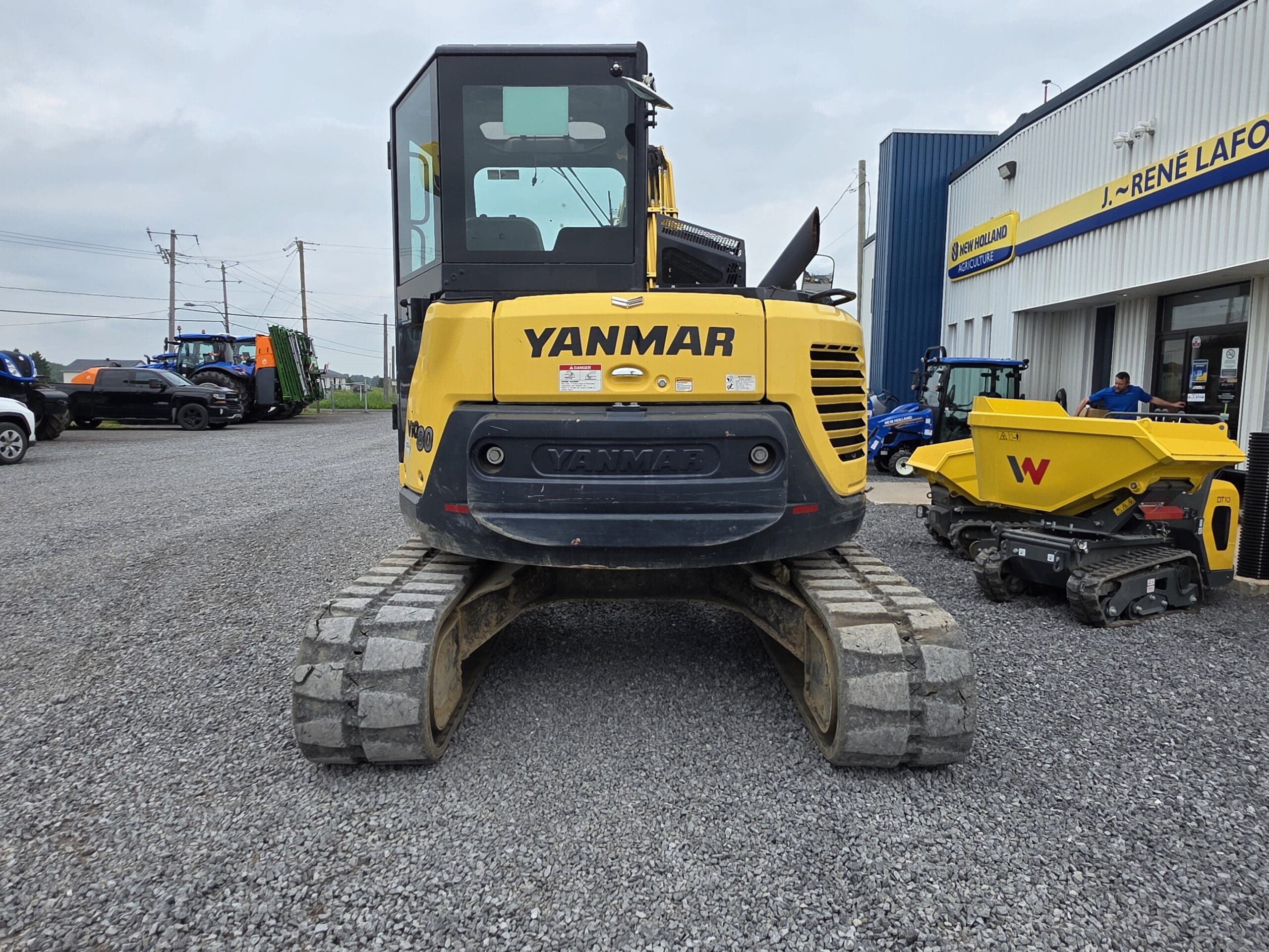 Une pelleteuse Yanmar jaune est garée sur du gravier devant un bâtiment bleu et blanc, avec d'autres véhicules de construction à proximité. La photo est prise de l'arrière de l'excavatrice.