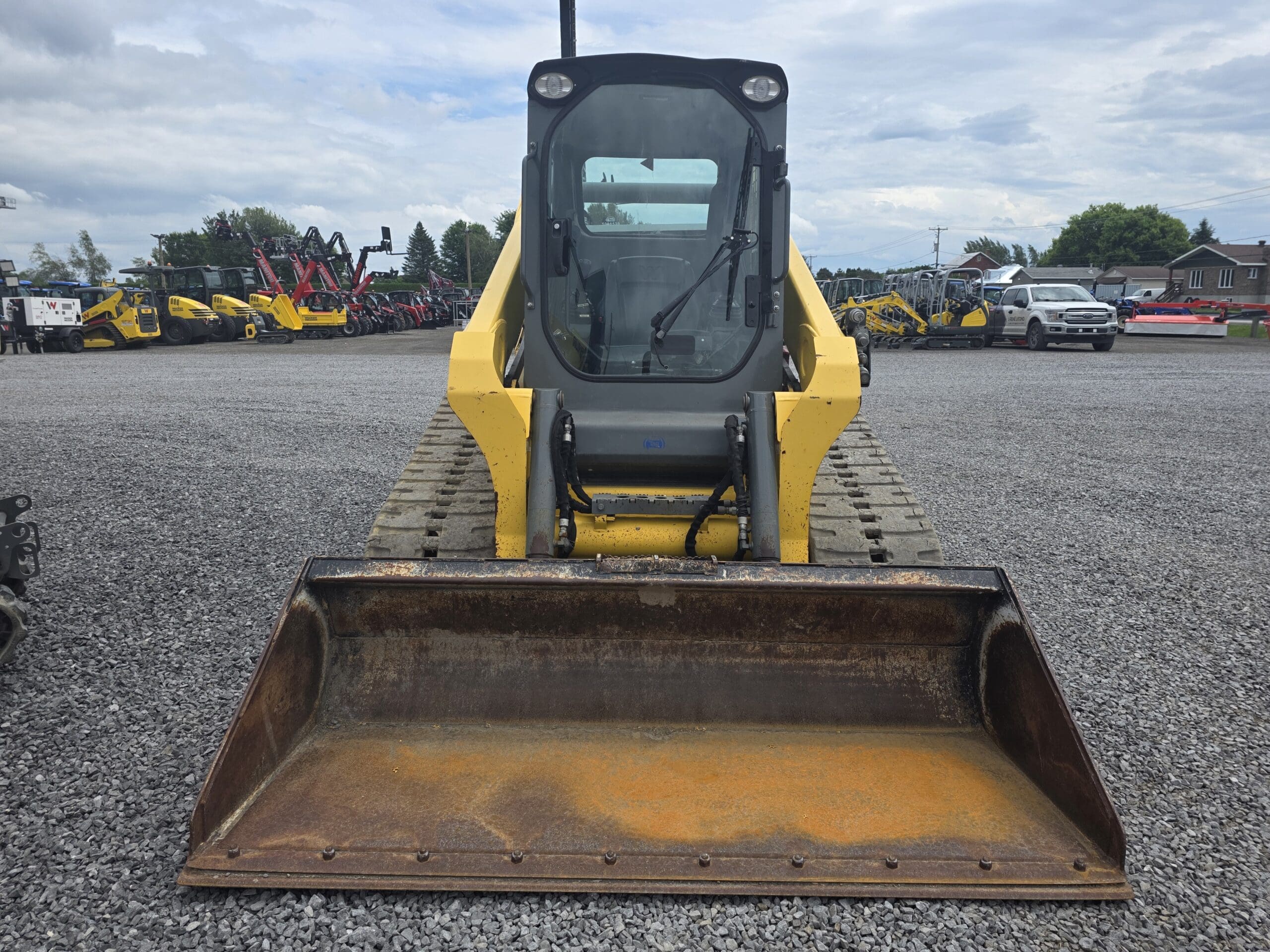 Une chargeuse compacte jaune avec un grand godet avant est garée sur un terrain en gravier, face à la caméra. D'autres véhicules et camions de construction sont visibles à l'arrière-plan sous un ciel nuageux.