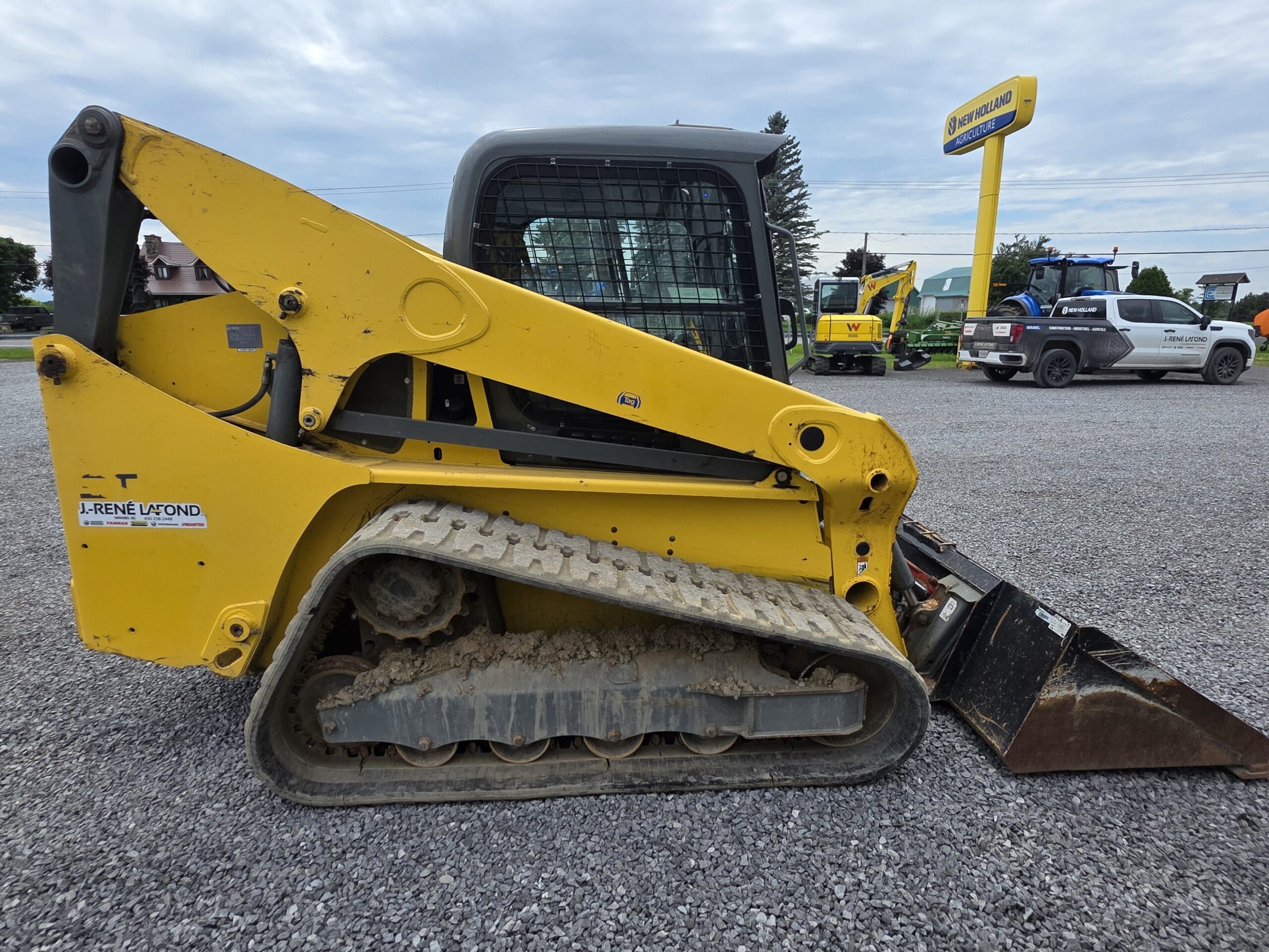 Une chargeuse compacte sur chenilles jaune, équipée d'un godet métallique, est garée sur du gravier. Plusieurs véhicules et engins de chantier sont visibles à l'arrière-plan par une journée nuageuse.