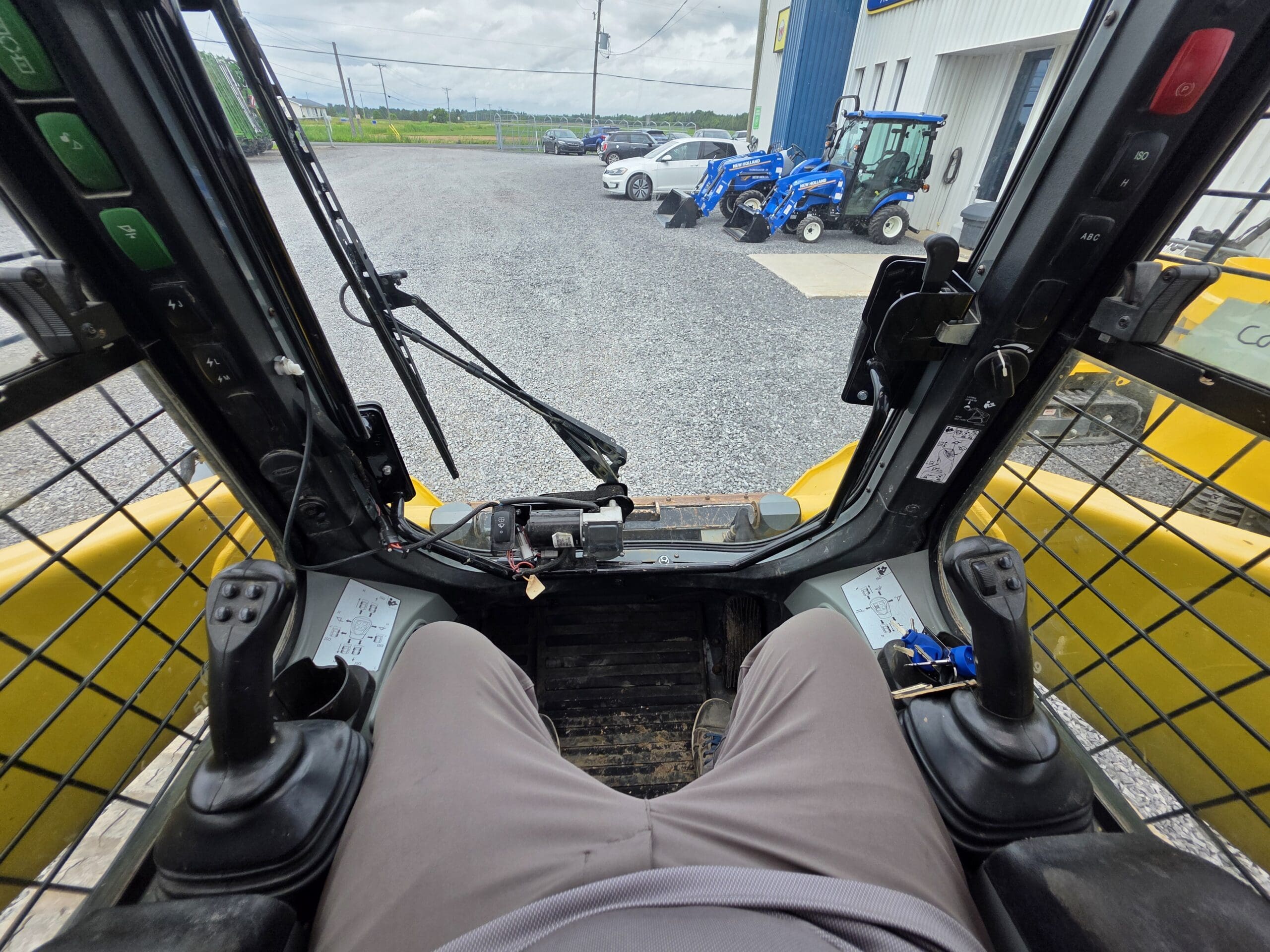 Vue de l'intérieur de la cabine d'un véhicule de chantier jaune, montrant les jambes de l'opérateur, les leviers de commande et la fenêtre. À l'extérieur, des tracteurs et des voitures sont garés sur un terrain en gravier près d'un bâtiment.