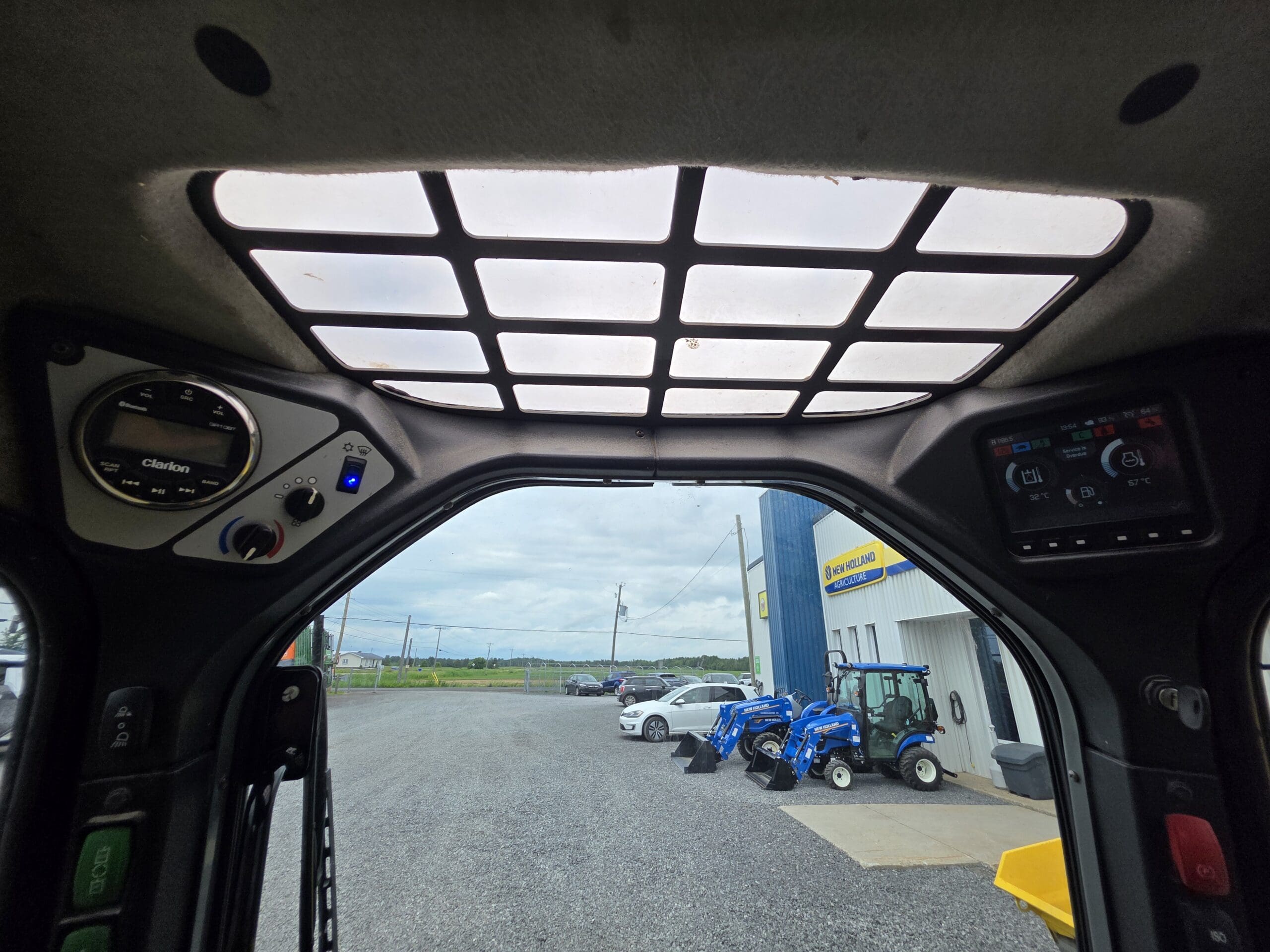 Vue de l'intérieur d'un véhicule, à travers une fenêtre de toit quadrillée. À l'extérieur, plusieurs tracteurs bleus sont garés près d'un bâtiment sur un terrain en gravier, sous un ciel nuageux. Les commandes des véhicules sont visibles au-dessus de la tête.