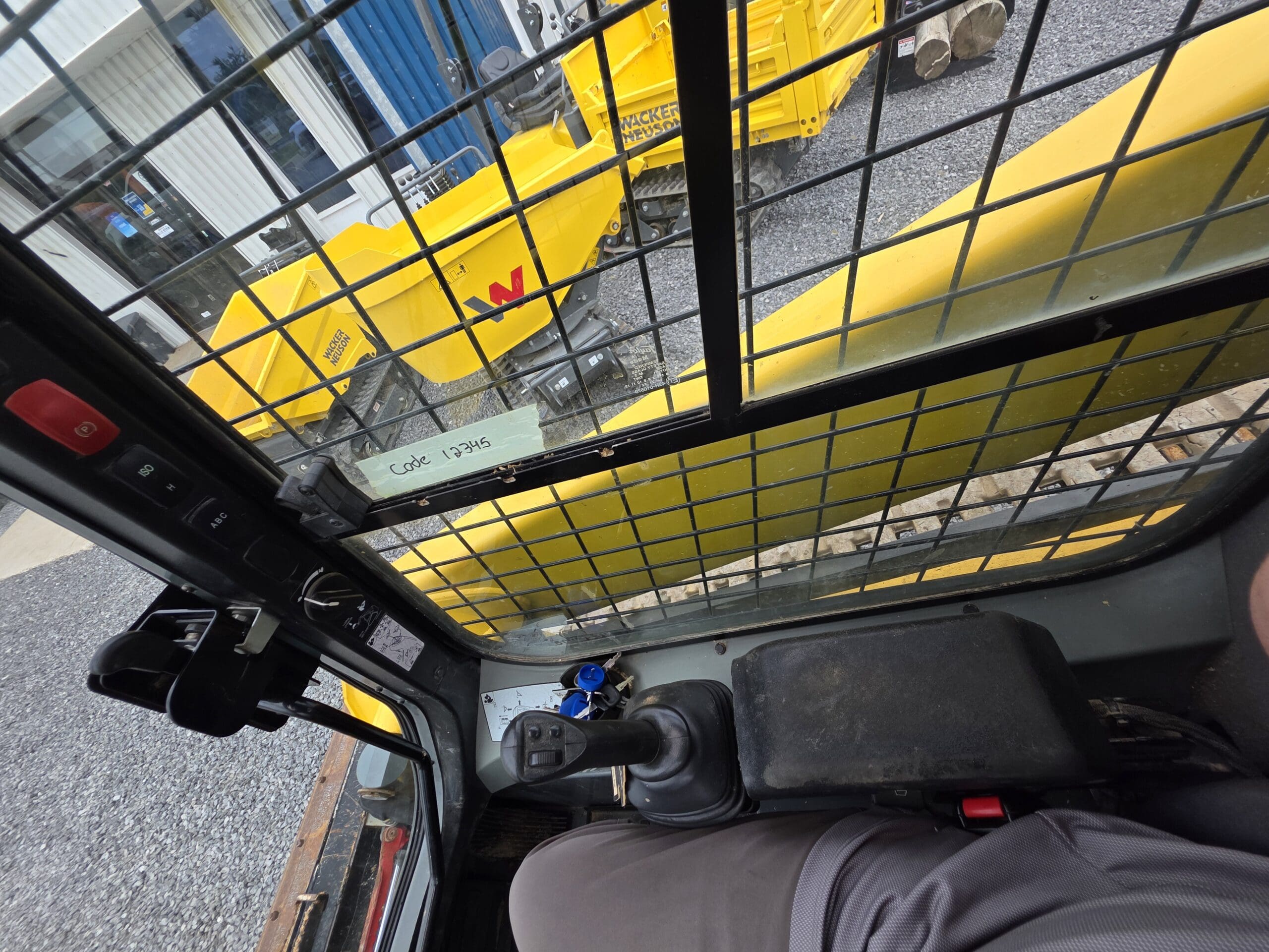 Vue de l'intérieur de la cabine d'un véhicule de construction, montrant le joystick de commande et le siège. À l'extérieur, une machine Wacker Neuson jaune et un terrain en gravier sont visibles à travers une fenêtre à grille métallique.