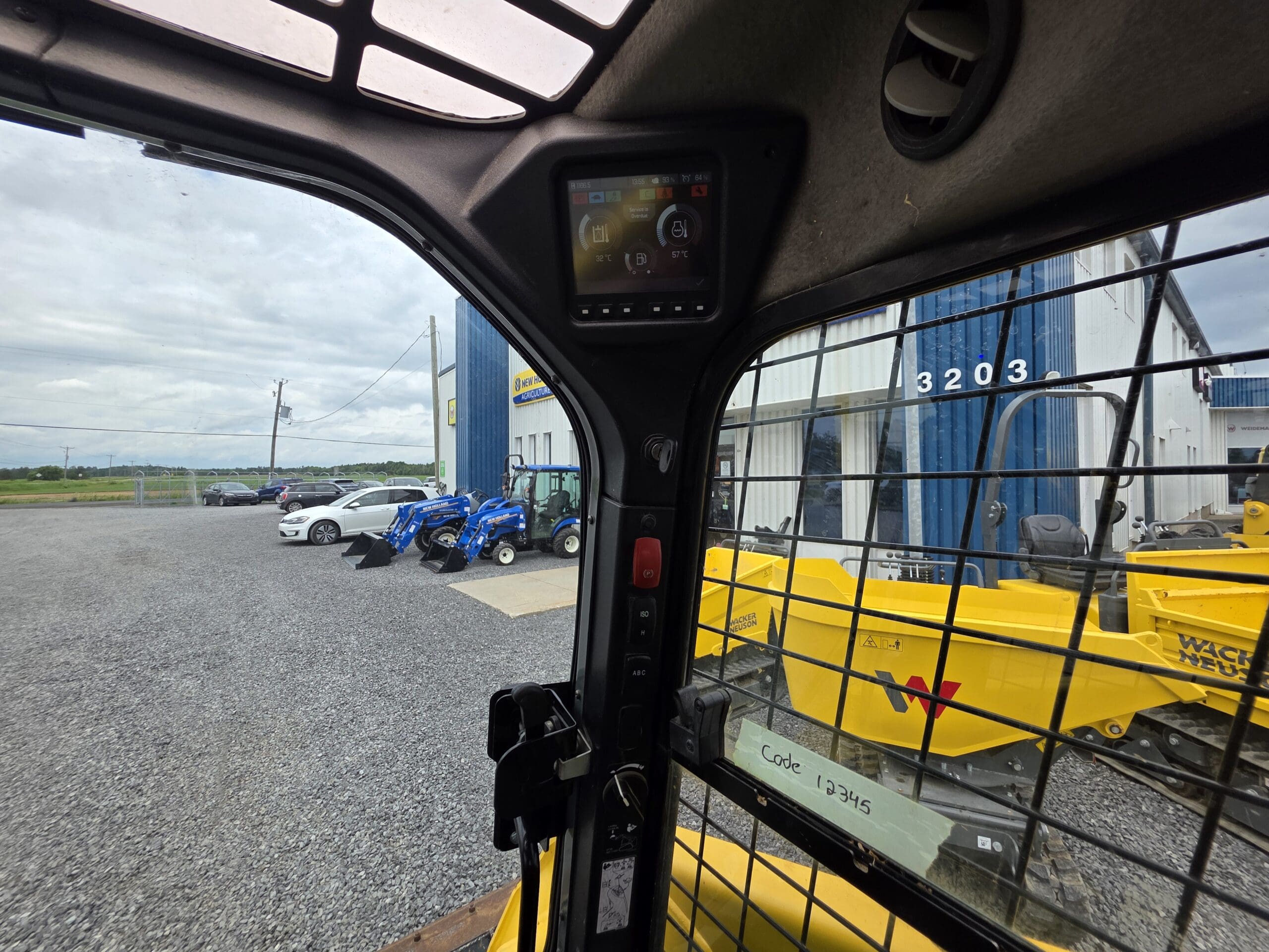 Vue de l'intérieur de la cabine d'un véhicule de chantier, à travers une fenêtre métallique grillagée, sur des machines bleues et jaunes et des voitures garées à l'extérieur d'un bâtiment numéroté 3203. Un panneau de commande est visible au-dessus de la fenêtre.
