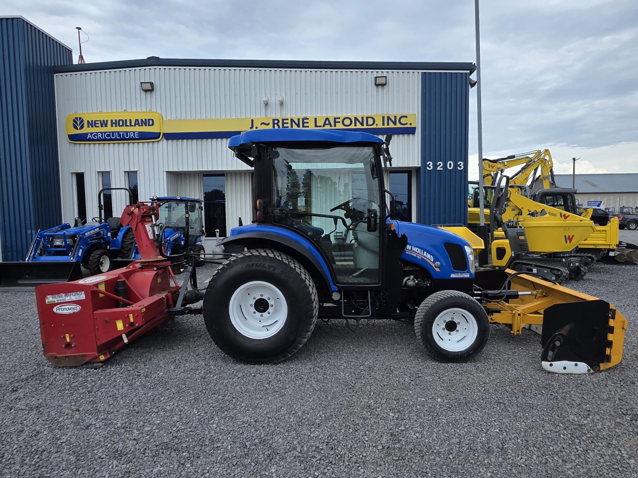 Un tracteur New Holland bleu avec des accessoires rouges et jaunes est garé devant un bâtiment portant l'inscription