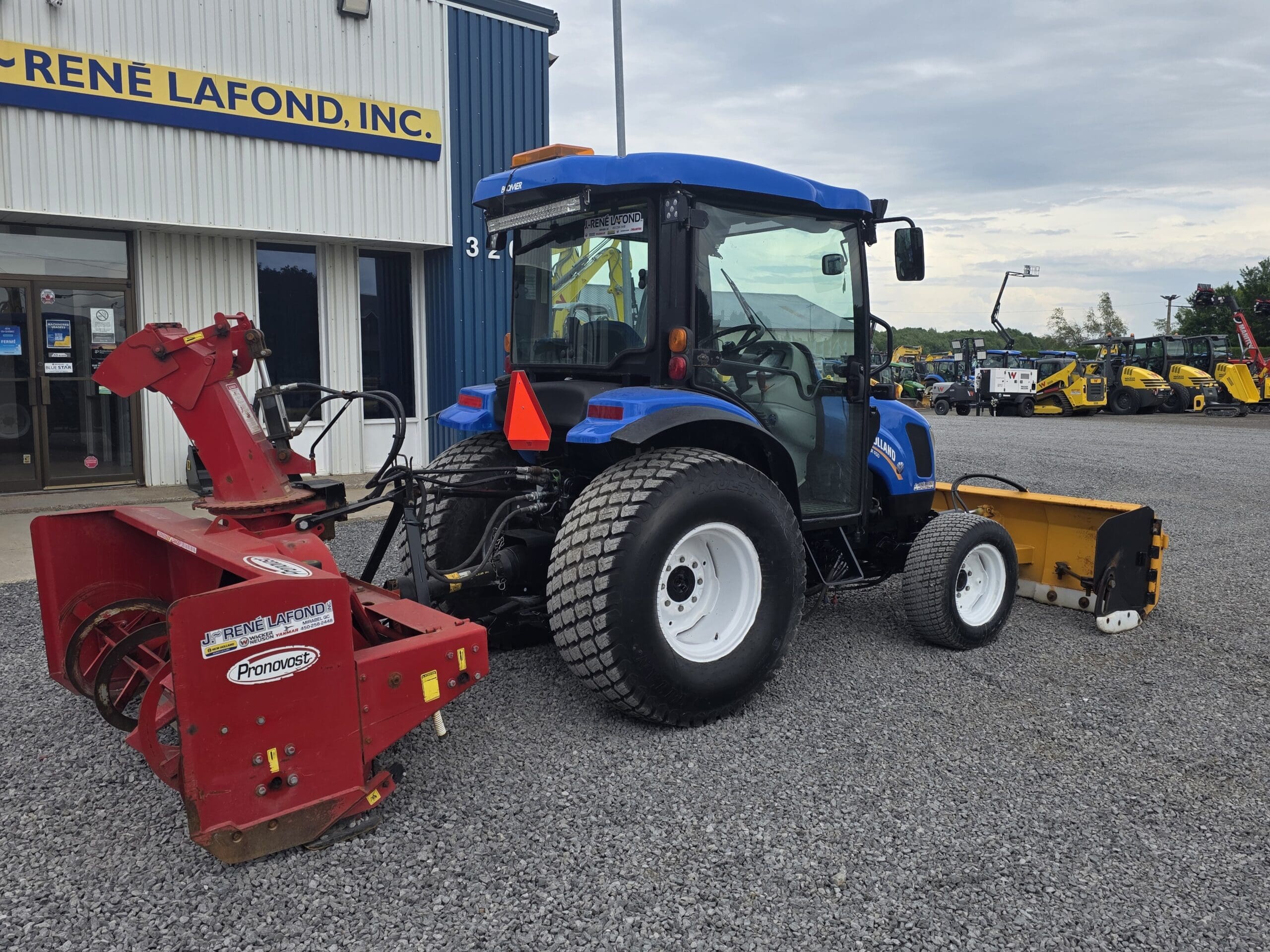 Un tracteur bleu équipé d'une souffleuse à neige rouge et d'un accessoire jaune est garé sur du gravier à l'extérieur d'un bâtiment portant l'inscription