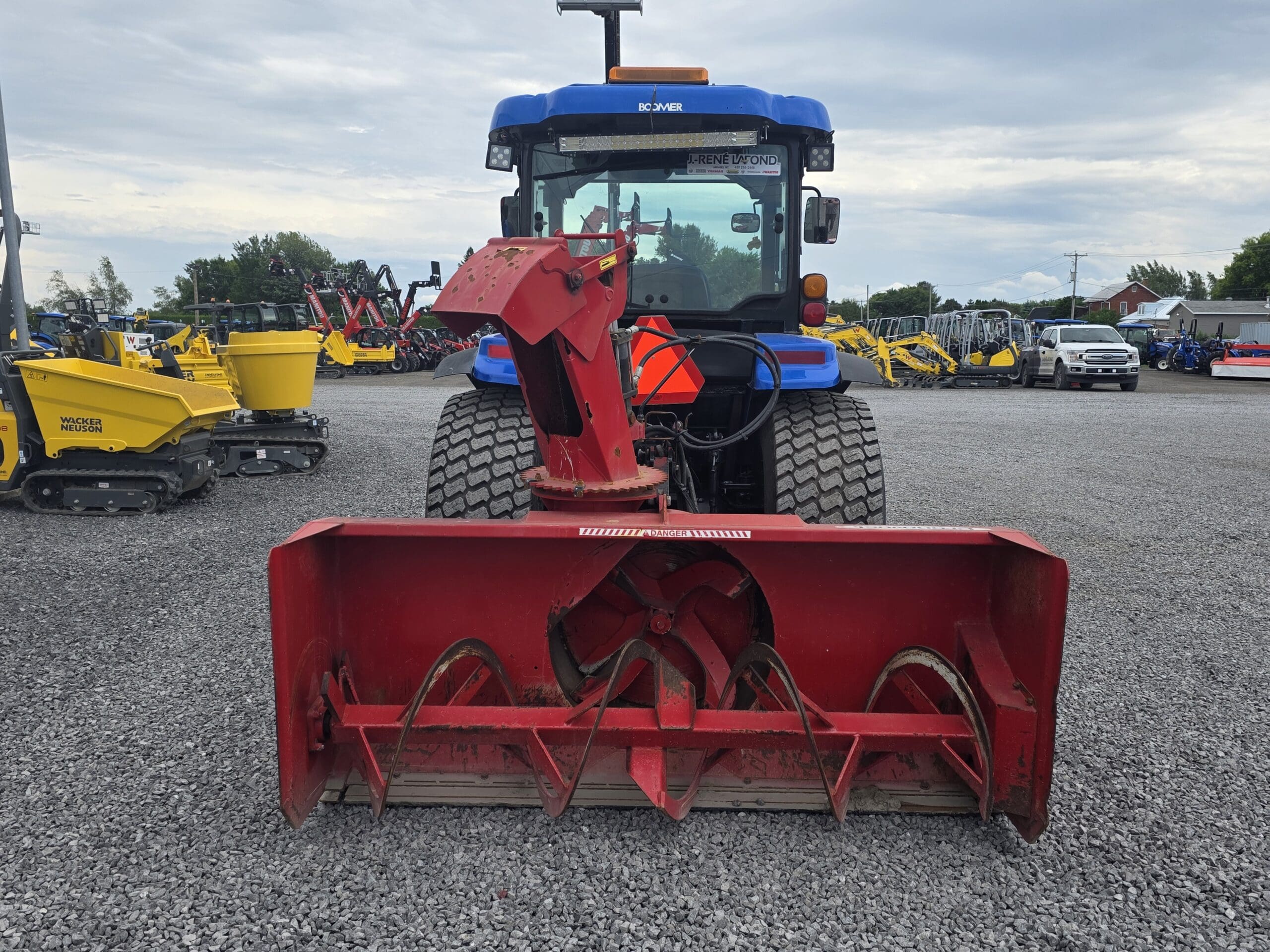 Un tracteur bleu à gros pneus est garé sur du gravier, vu de l'avant. À l'avant se trouve une souffleuse à neige rouge avec des tarières visibles. Des engins de chantier sont visibles à l'arrière-plan.