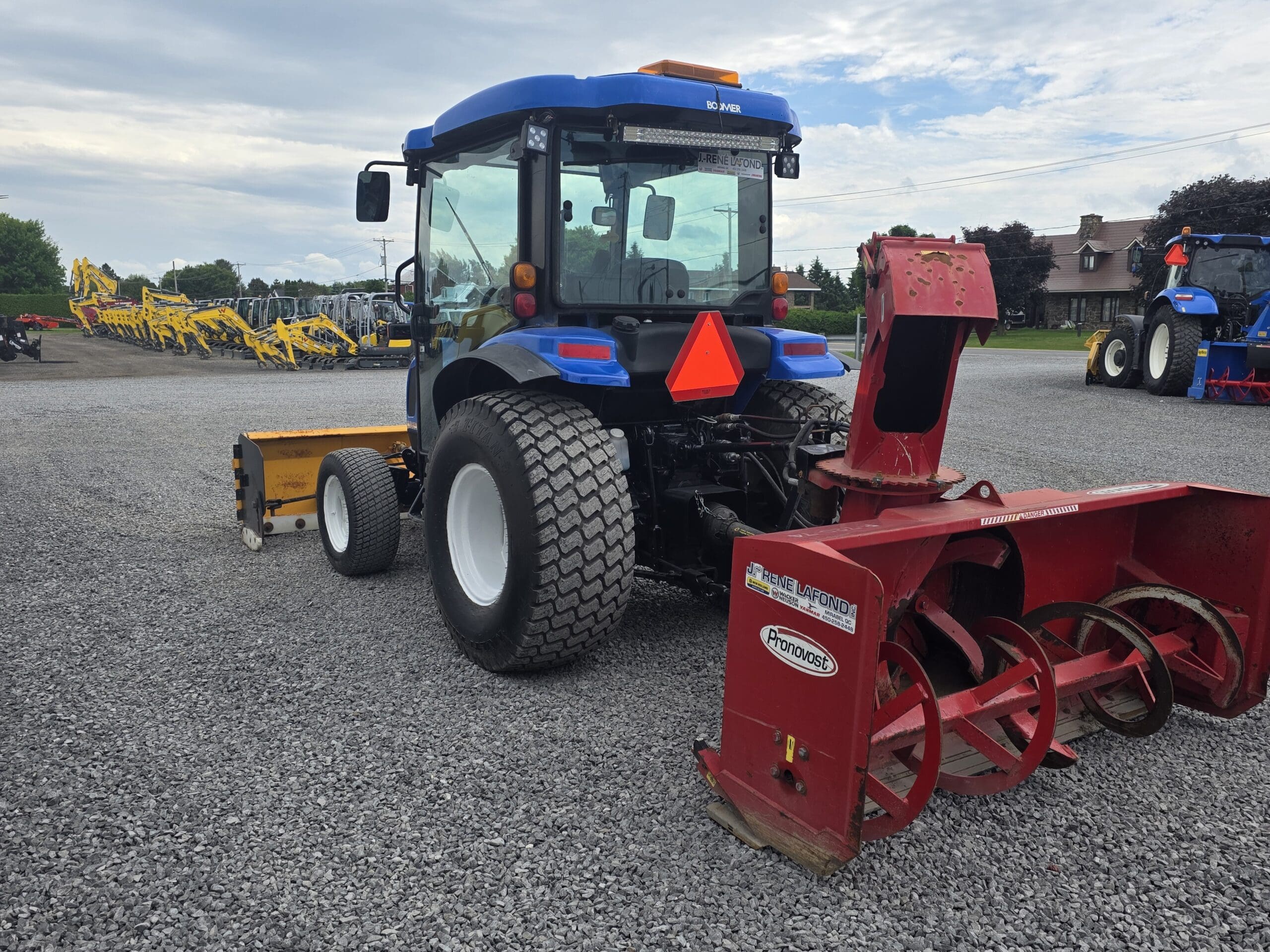 Un tracteur bleu avec de gros pneus arrière est garé sur du gravier, attelé à une souffleuse à neige rouge et à un outil jaune. D'autres machines et une maison sont visibles à l'arrière-plan sous un ciel partiellement nuageux.