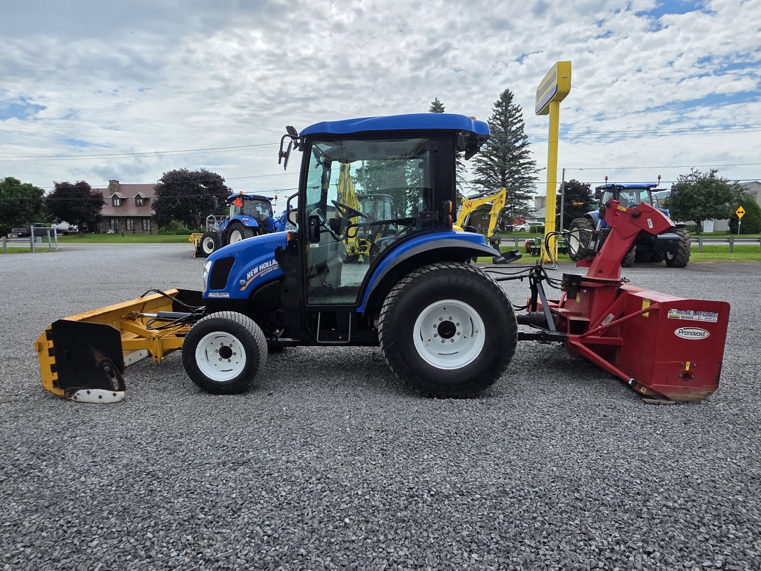 Un tracteur bleu équipé d'un outil avant jaune et d'un grand outil arrière rouge est garé sur un terrain en gravier, avec des arbres, des bâtiments et d'autres tracteurs visibles en arrière-plan, sous un ciel partiellement nuageux.