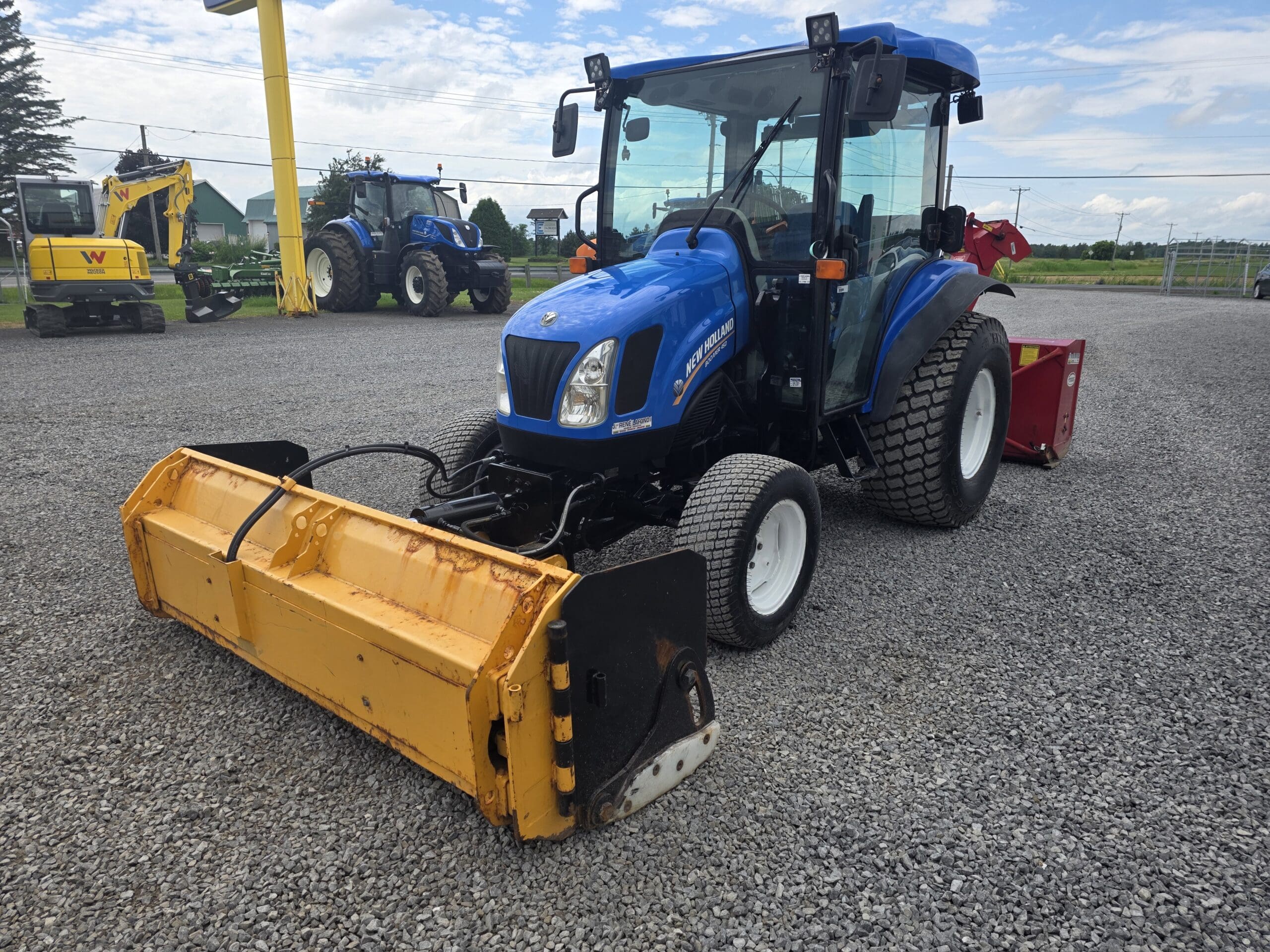 Un tracteur New Holland Boomer 50 bleu avec un accessoire jaune à l'avant est garé sur du gravier, avec d'autres équipements agricoles et tracteurs visibles à l'arrière-plan.
