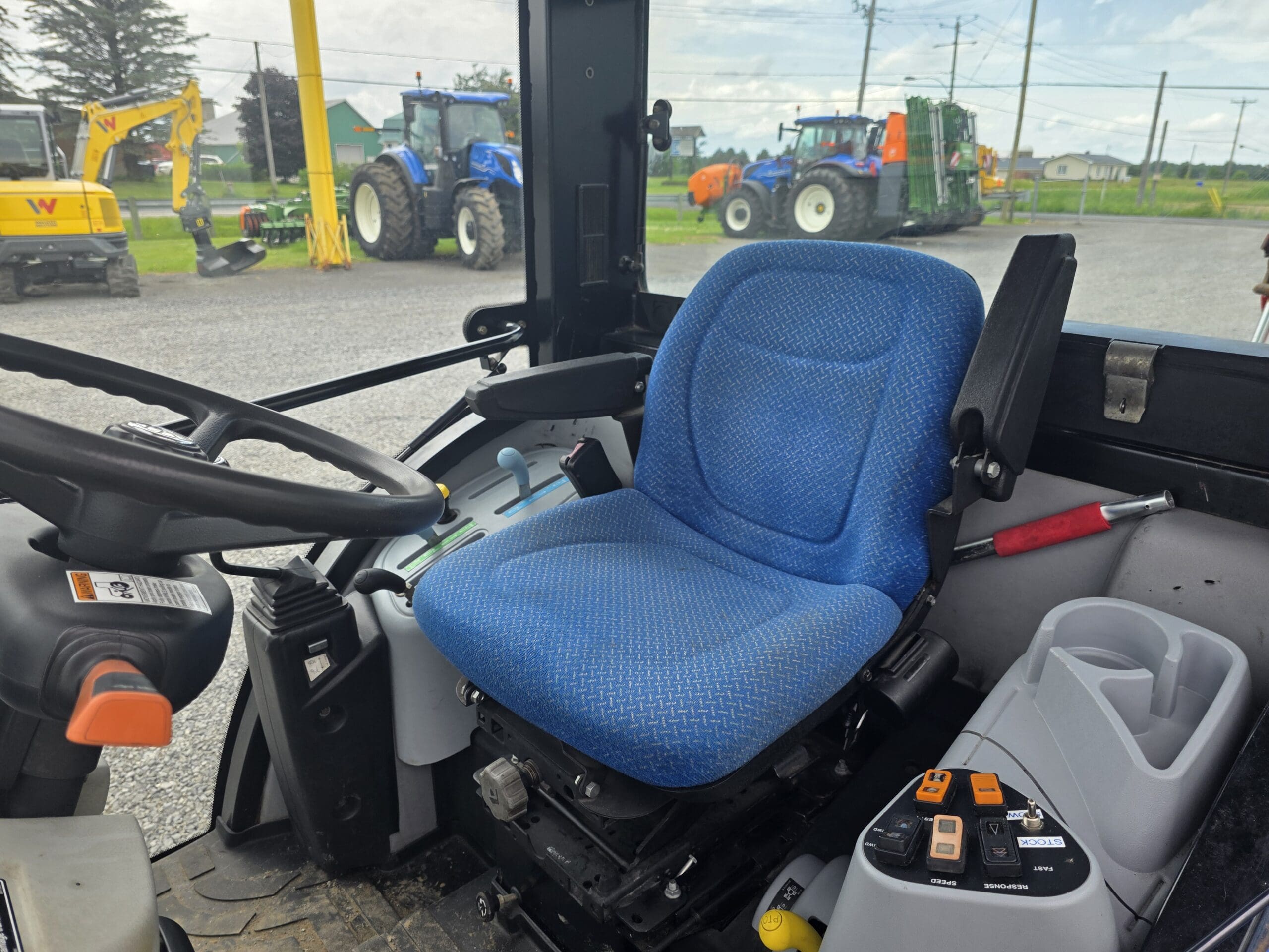 Intérieur d'une cabine de tracteur montrant un siège de conducteur bleu, un volant, divers boutons et leviers de commande, et de grandes fenêtres avec du matériel agricole et des véhicules visibles à l'extérieur.