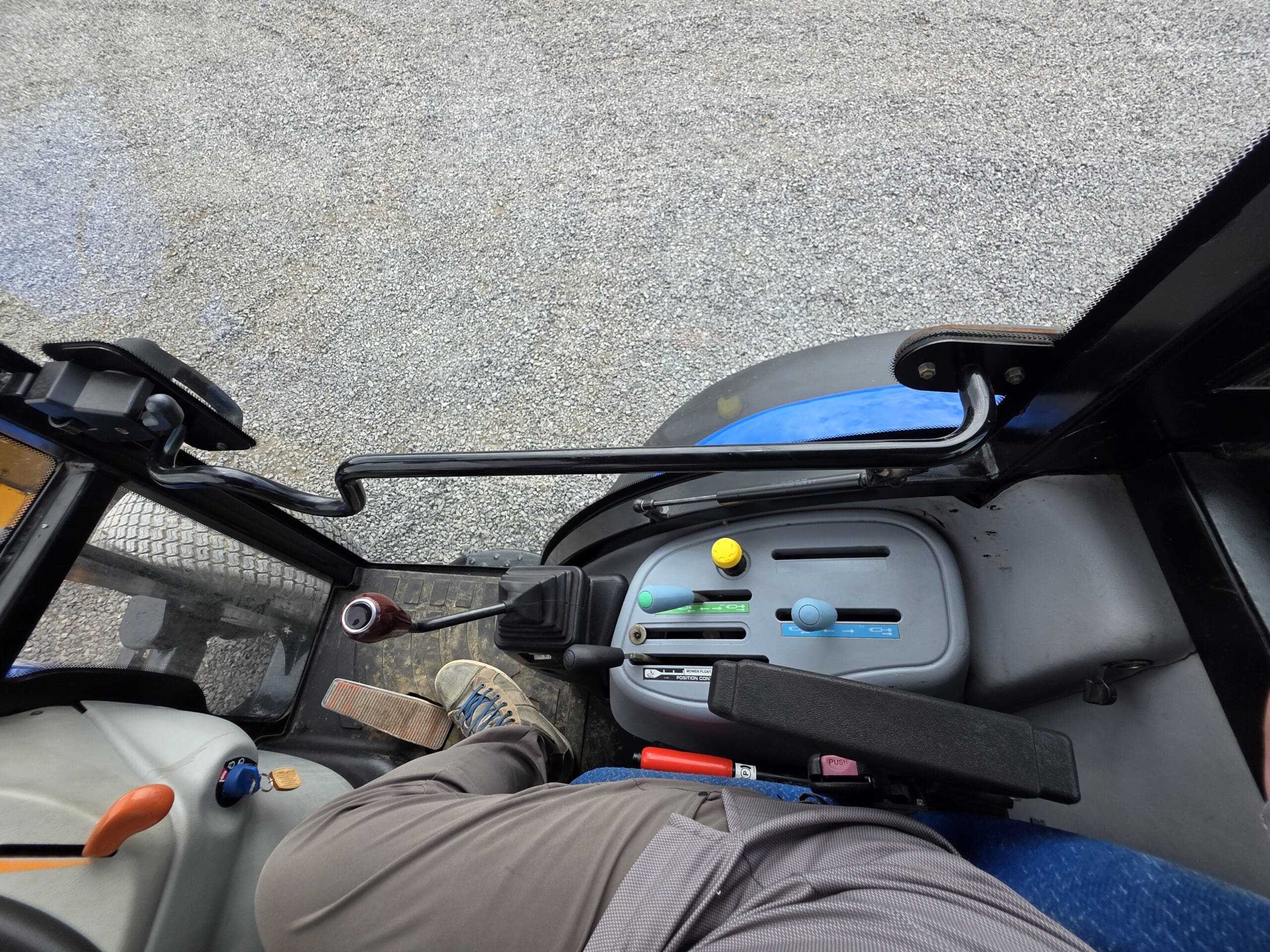 Vue de dessus de la cabine d'un tracteur montrant la jambe d'une personne, les commandes de vitesse avec des boutons colorés, les pédales et une partie du volant, avec une surface de gravier visible à l'extérieur de la fenêtre.