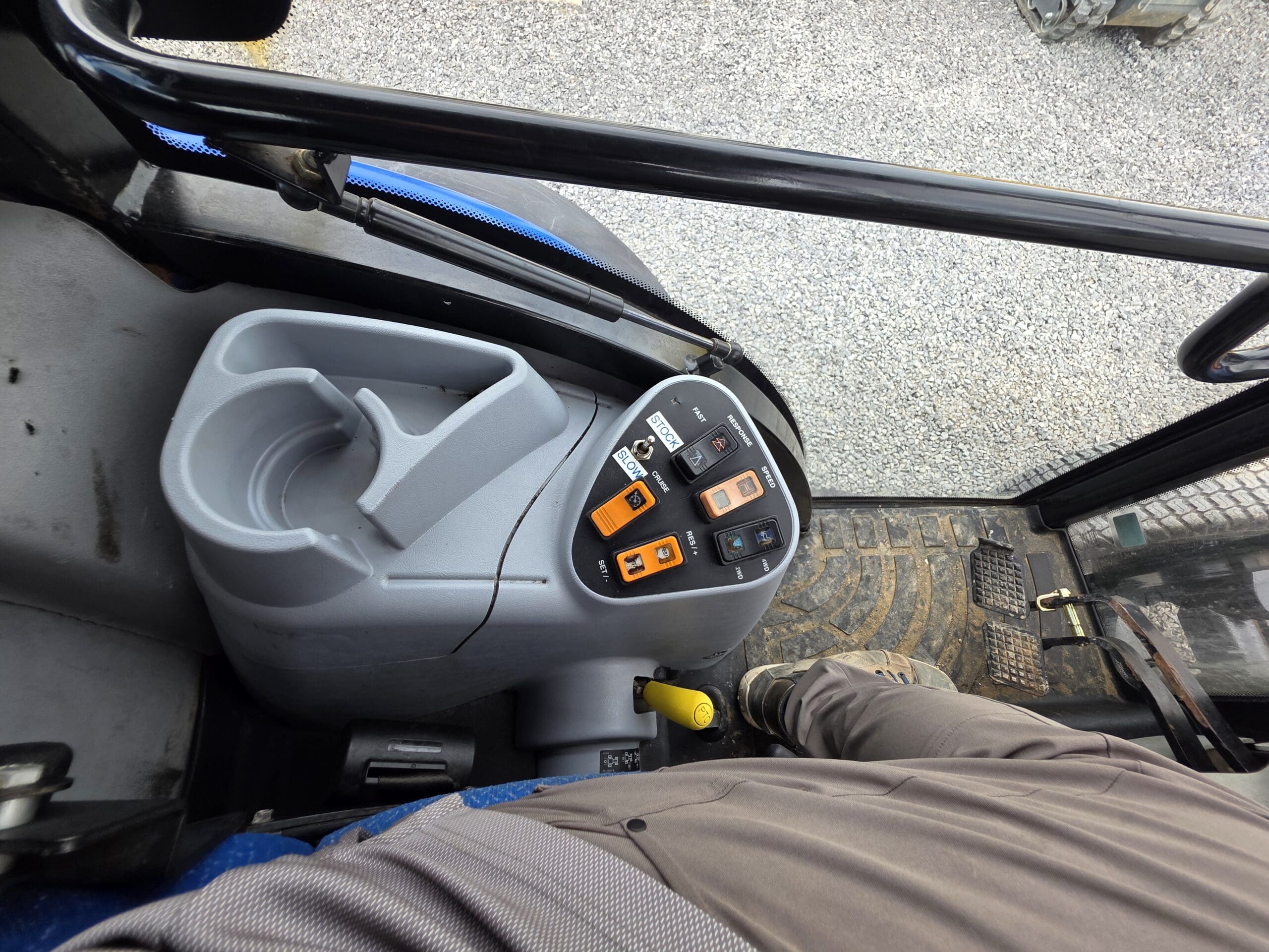 Vue de dessus à l'intérieur d'une cabine de véhicule montrant des boutons de commande et des interrupteurs à côté d'un porte-gobelet, de la jambe d'une personne et d'une partie d'un tapis de sol texturé avec des graviers visibles à l'extérieur par une fenêtre.