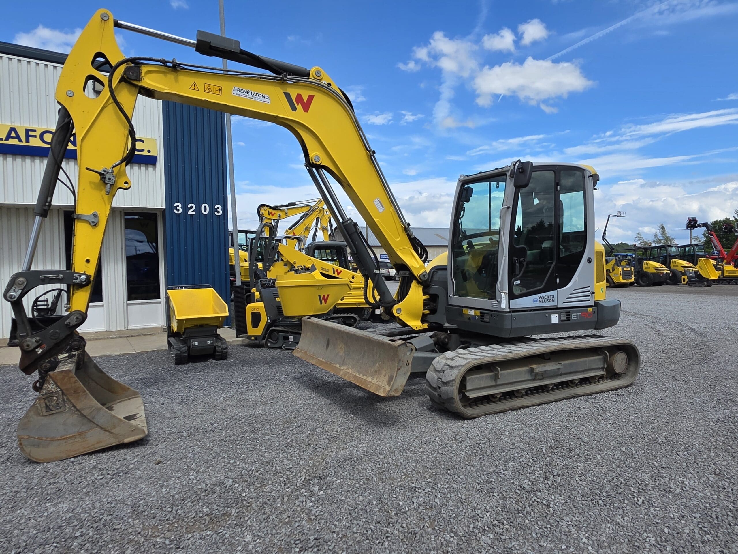 Une pelleteuse compacte jaune et grise avec un godet est garée sur du gravier à l'extérieur d'un bâtiment, avec d'autres équipements de construction visibles à l'arrière-plan sous un ciel partiellement nuageux.