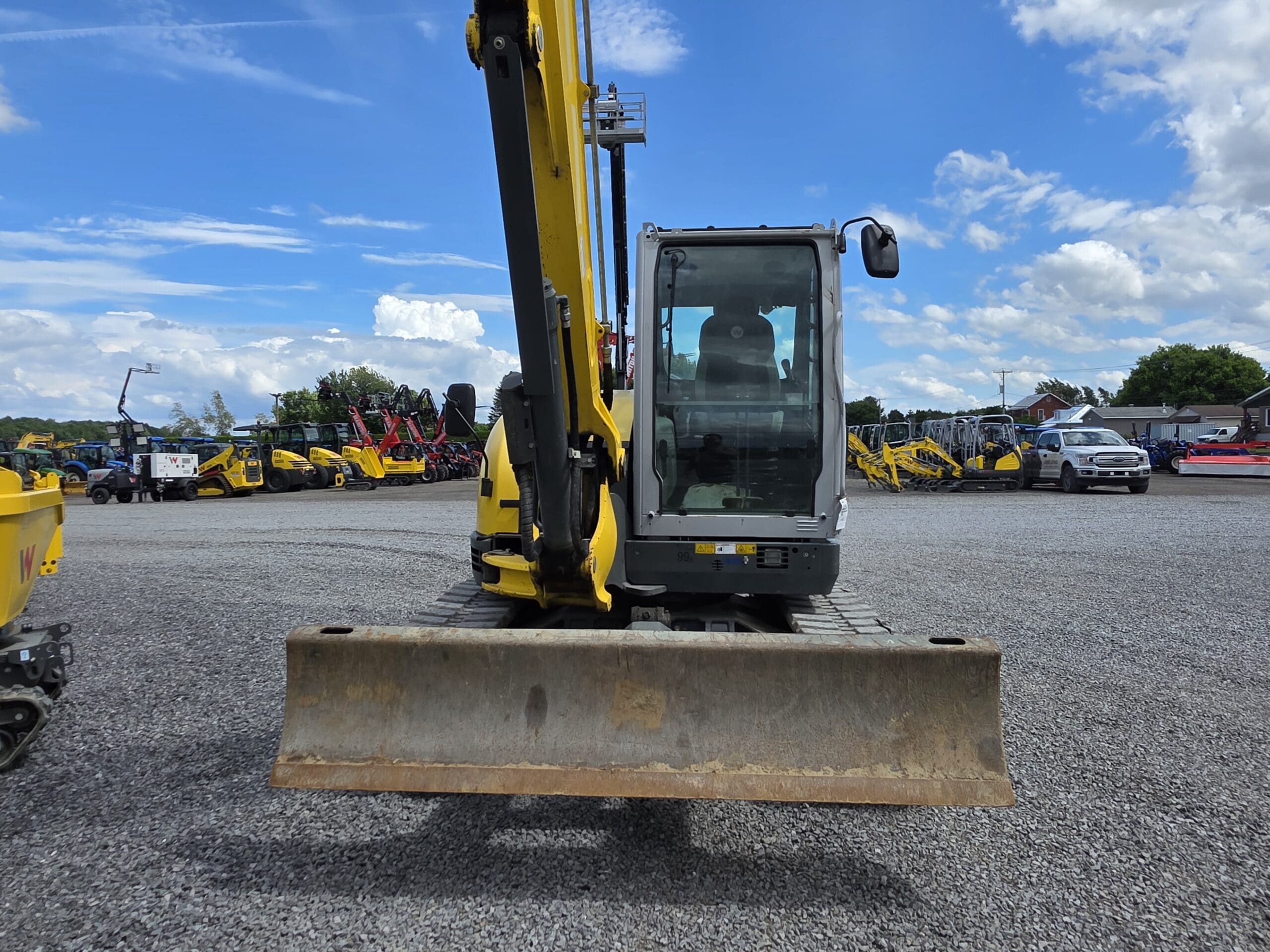 Une pelleteuse jaune avec une grande lame avant est garée sur un terrain en gravier, entourée de divers véhicules de construction, sous un ciel bleu partiellement nuageux.