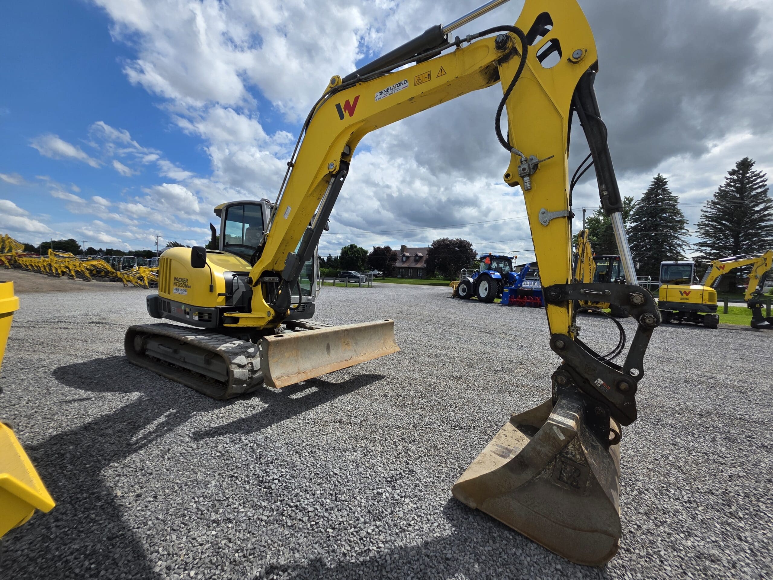Une pelleteuse jaune équipée d'un godet d'excavation est garée sur un terrain en gravier sous un ciel partiellement nuageux, avec d'autres équipements de construction visibles à l'arrière-plan.