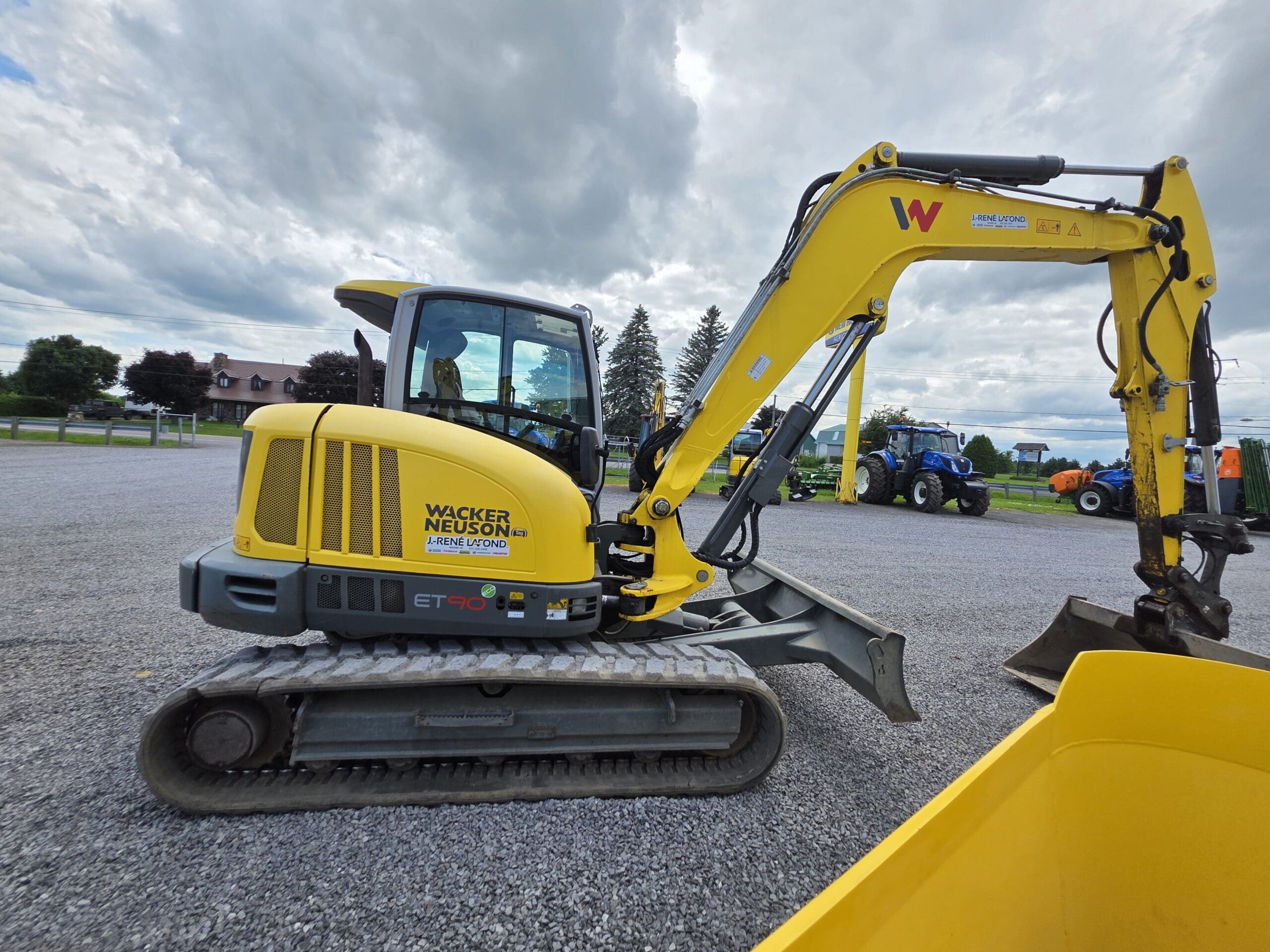 Une pelle compacte Wacker Neuson ET90 jaune garée sur du gravier, avec des arbres, une maison et des tracteurs en arrière-plan sous un ciel nuageux.