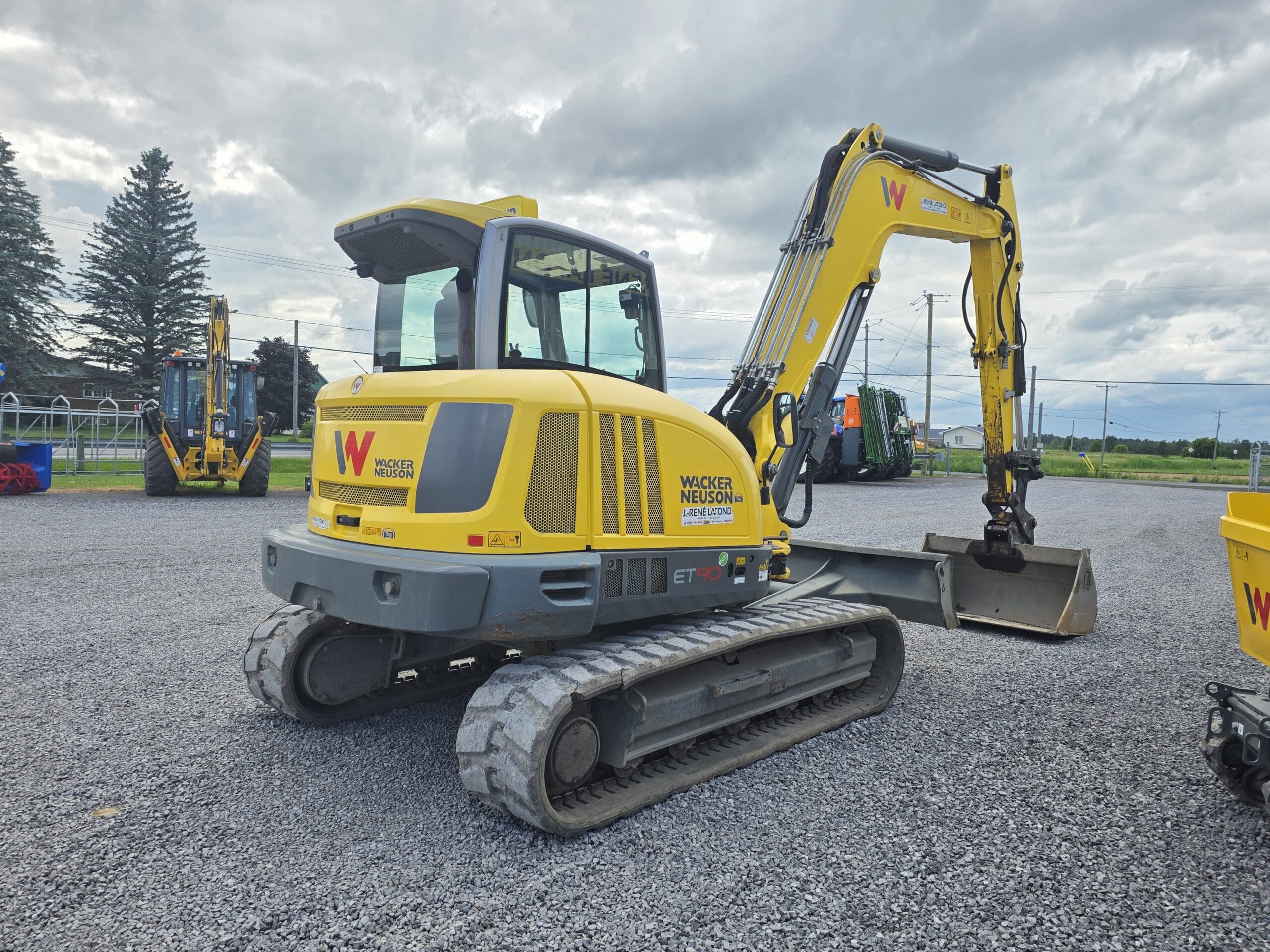 Une pelle compacte Wacker Neuson jaune avec un godet d'excavation est garée sur du gravier avec d'autres équipements de construction et des arbres en arrière-plan sous un ciel nuageux.