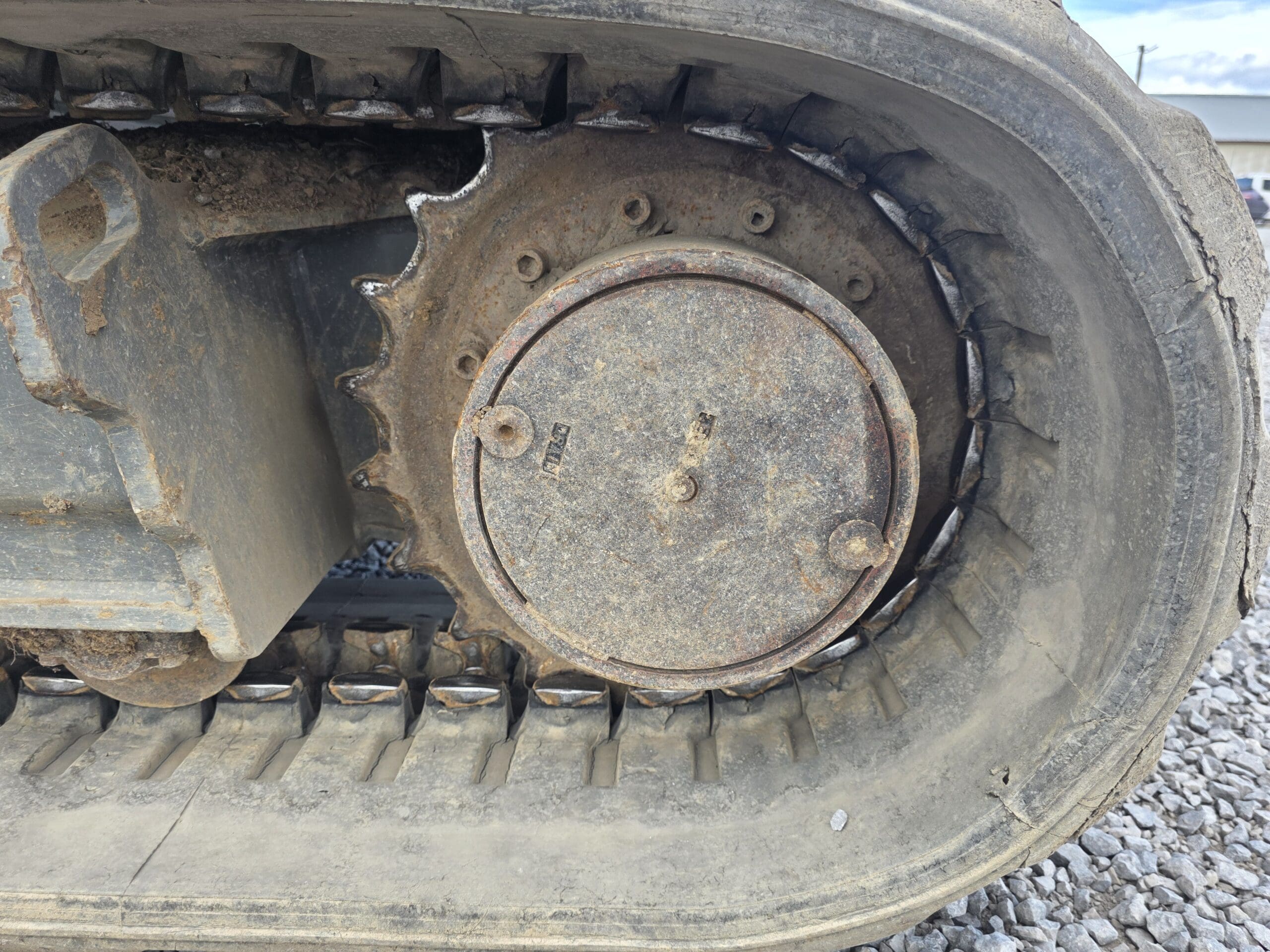 Vue rapprochée d'une chenille métallique et d'une roue dentée sur un véhicule de construction, tel qu'une pelleteuse ou un bulldozer, avec de la saleté et de la rouille visibles ; du gravier se trouve sur le sol à l'arrière-plan.