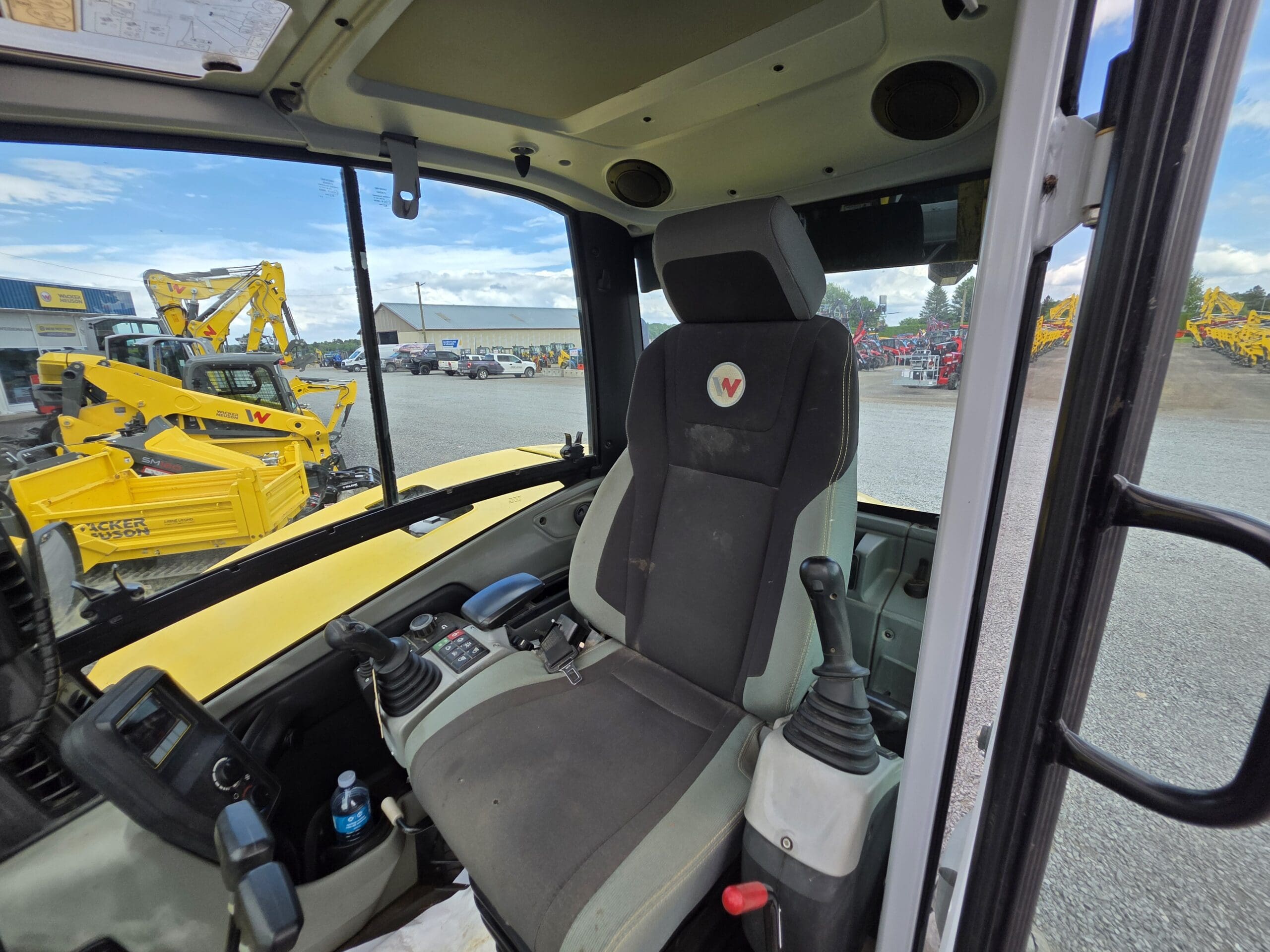 Vue de l'intérieur de la cabine d'un véhicule de chantier, montrant le siège du conducteur, le volant, les commandes et une fenêtre avec divers équipements de chantier jaunes visibles à l'extérieur.
