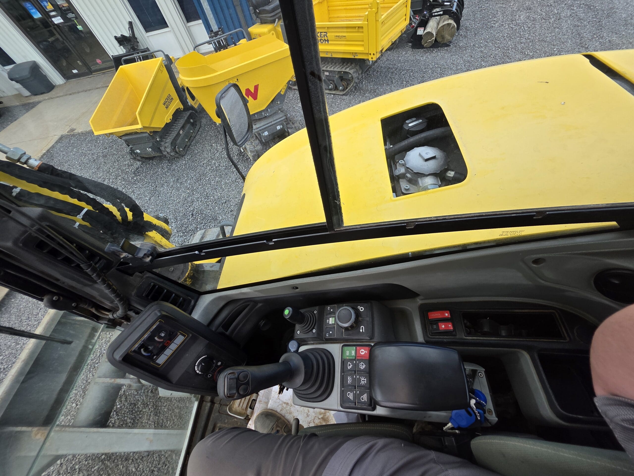 Vue de l'intérieur de la cabine d'un véhicule de chantier jaune, montrant des panneaux de commande, des manettes et une partie de la jambe d'une personne. À l'extérieur, deux chariots utilitaires jaunes et un sol en gravier sont visibles à travers les fenêtres.