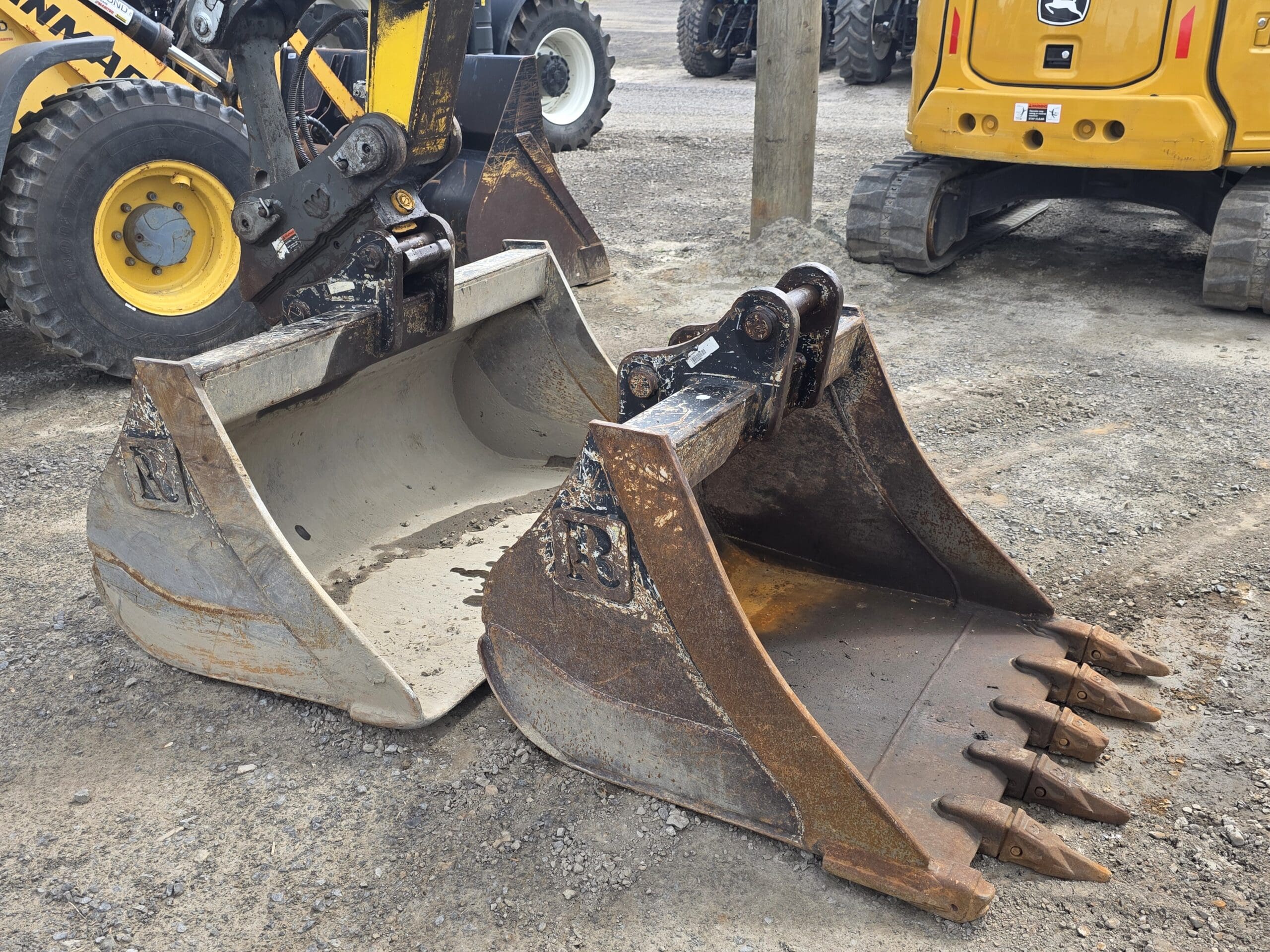 Deux grands godets de pelleteuse, l'un lisse et gris, l'autre rouillé avec des dents pointues, sont attachés à des véhicules de construction jaunes, posés sur un terrain en gravier.