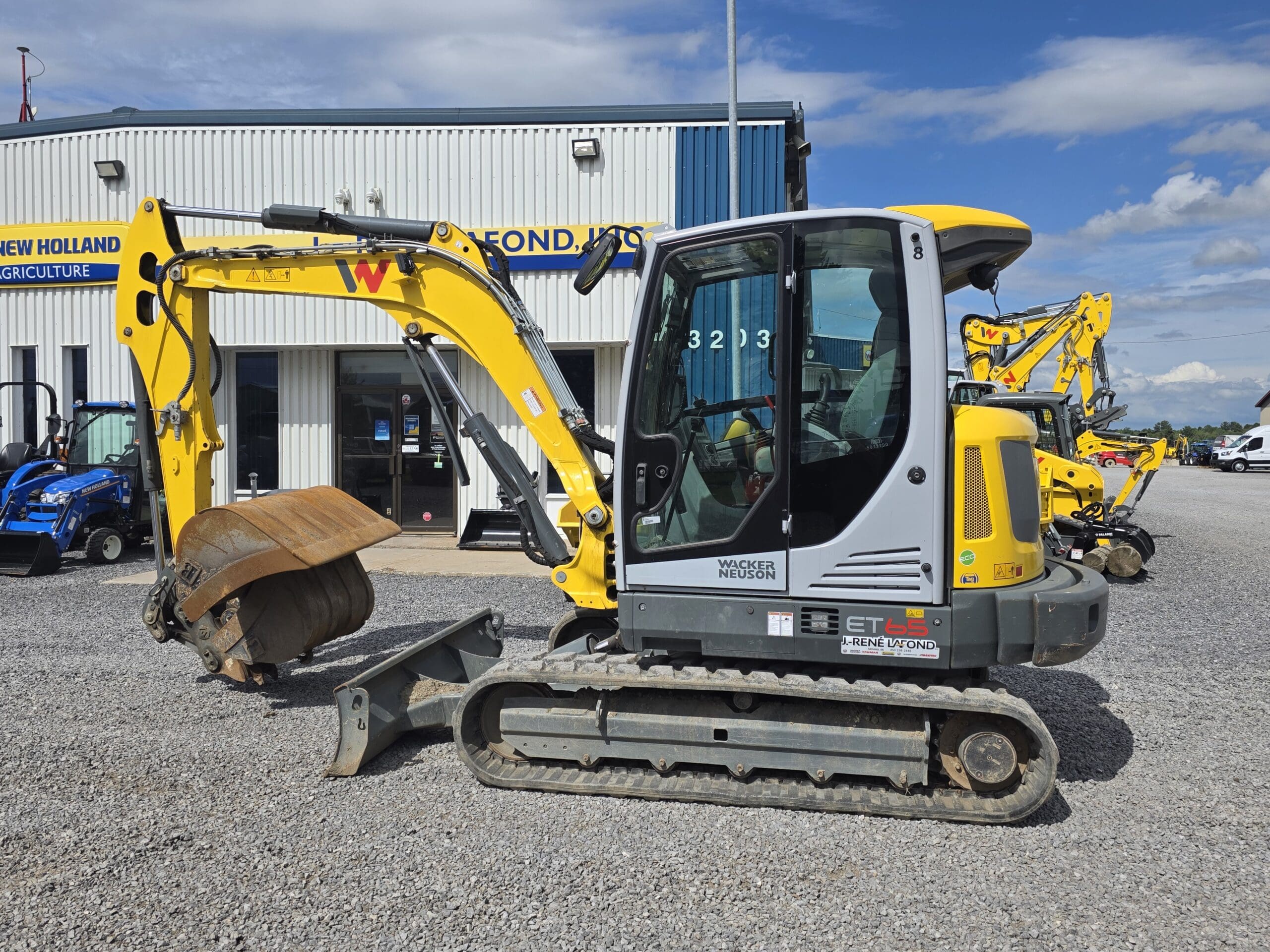 Une mini-pelle jaune et grise est garée sur un terrain en gravier devant le bâtiment d'un concessionnaire New Holland, sous un ciel bleu avec quelques nuages. D'autres véhicules de construction sont visibles à l'arrière-plan.