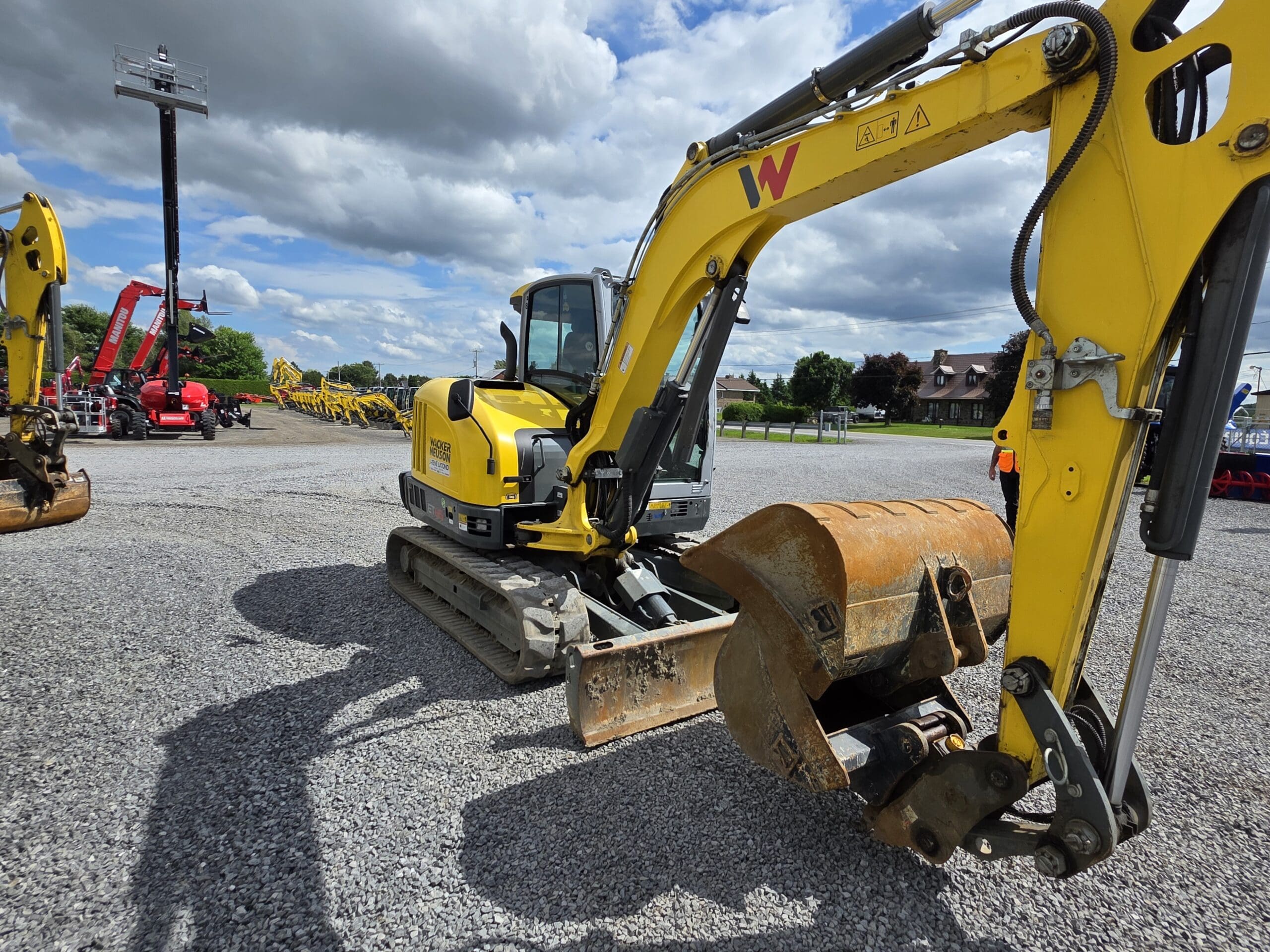 Une pelleteuse jaune munie d'un grand godet est garée sur du gravier sous un ciel partiellement nuageux. D'autres véhicules et équipements de construction sont visibles à l'arrière-plan.