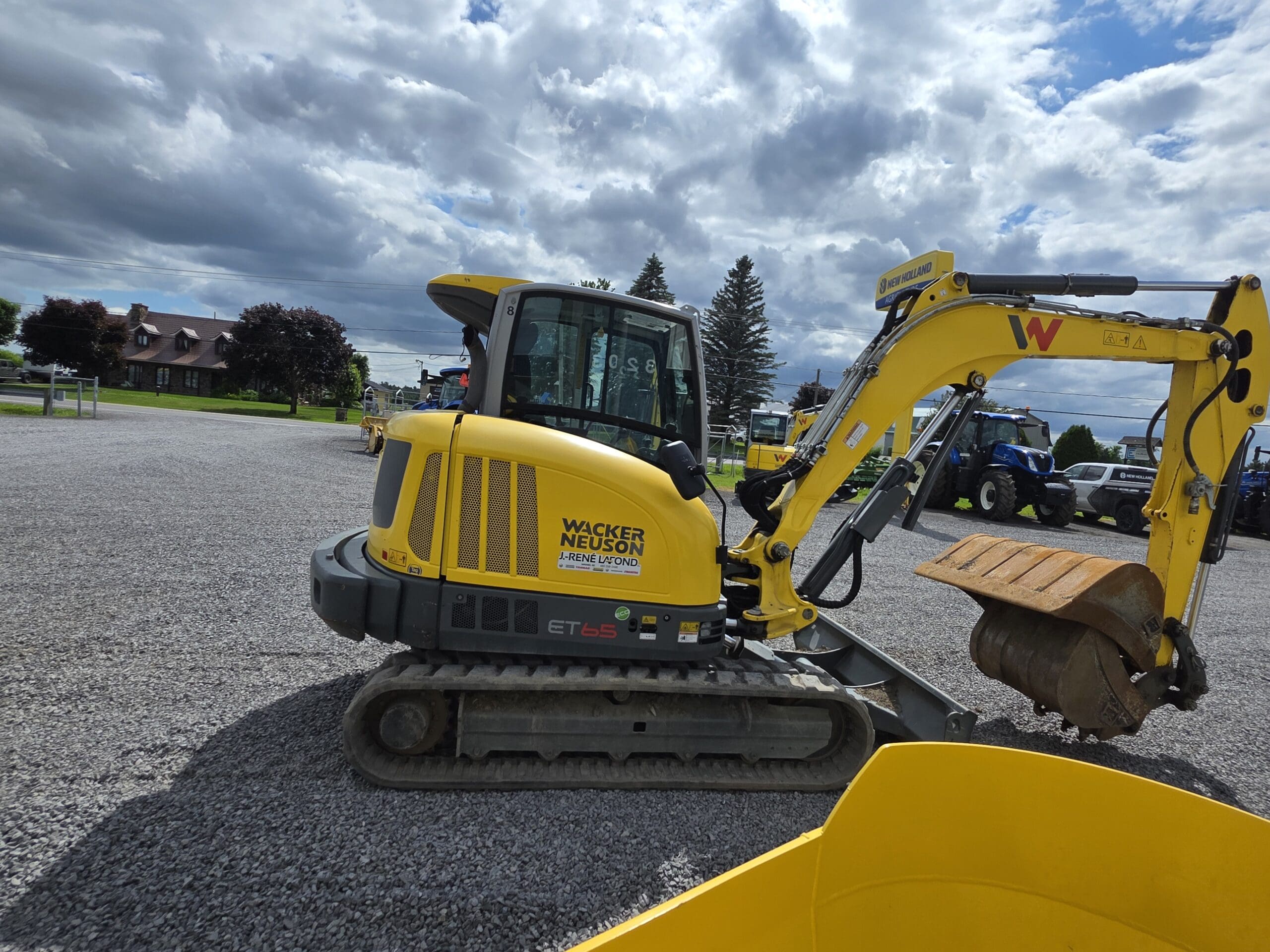 Une pelle compacte Wacker Neuson ET65 jaune est garée sur un terrain en gravier sous un ciel partiellement nuageux, avec des arbres, de l'herbe et d'autres machines visibles à l'arrière-plan.